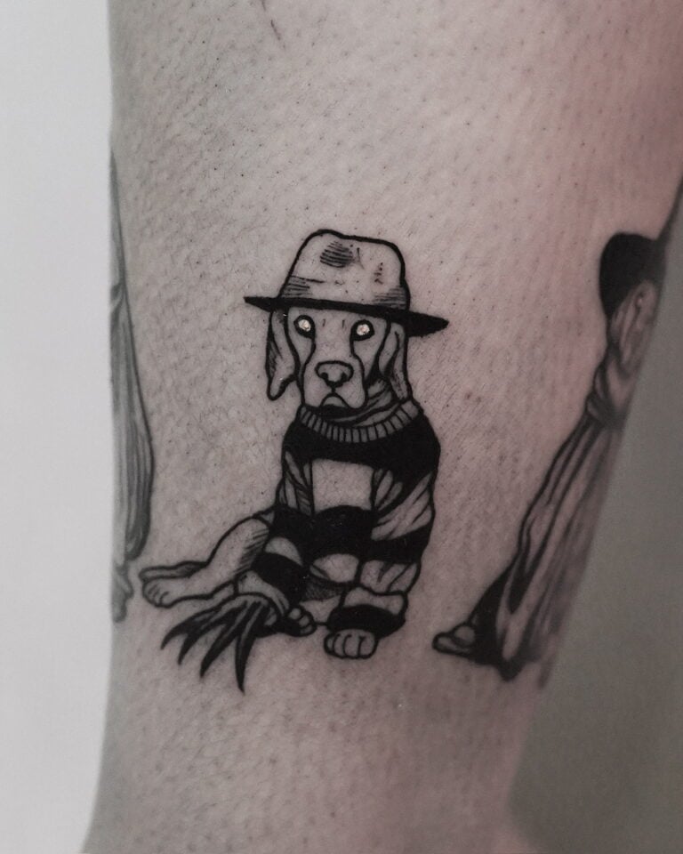 7sinstattoo.live Czarno-biały tatuaż, wykonany w studio tatuaży Poznań, przedstawia psa w szerokim kapeluszu i pasiastym swetrze, przypominającym postać z horroru. Pies ma pazury w rękawiczkach na jednej łapie, co dodaje niepokojącego motywu, i siedzi na jasnym ramieniu osoby. Tatuaż jest skatalogowany jako produkt DSC09972. Najlepsze studio tatuażu Seven Sins Tattoo