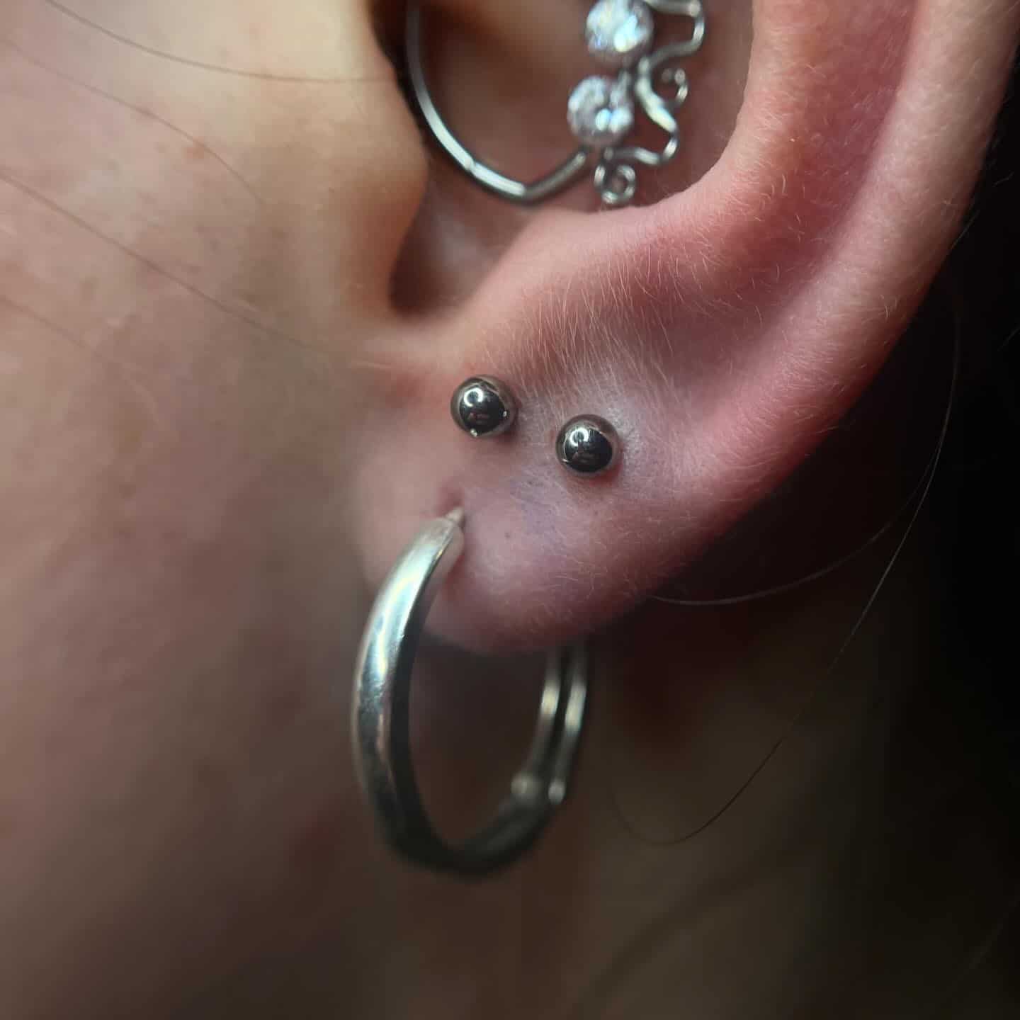 7sinstattoo.live Zbliżenie na ucho osoby z kilkoma piercingami, prezentowane w najlepszym studio tatuażu Poznań. W płatku ucha znajduje się duży srebrny pierścień, a w górnej chrząstce dwa małe kolczyki z czarnymi kamieniami szlachetnymi. W jednym z piercingów chrząstki widoczny jest również ozdobny srebrny element z dwoma małymi kryształkami z produktu E21971E2-9B81-481B-9AE2-0824CF6EB4D1. Najlepsze studio tatuażu Seven Sins Tattoo