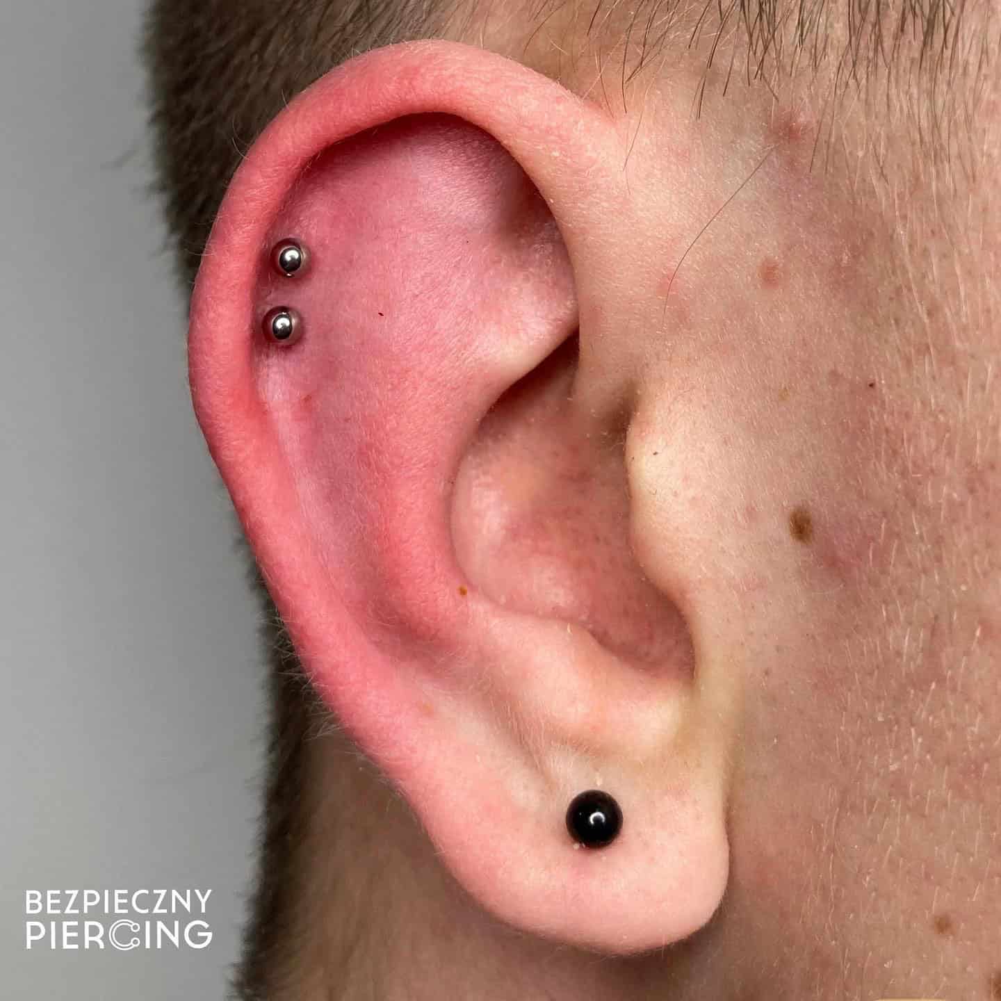 7sinstattoo.live Zbliżenie ucha z dwoma piercingami: jeden w górnej części ucha z dwoma metalowymi kolczykami i drugi w płatku ucha z jednym czarnym kolczykiem. Skóra wokół ucha wydaje się gładka i zdrowa. „E79536A4-026C-44F3-BBFA-6E295AA29393” autorstwa najlepsze studio tatuażu Poznań znajduje się w lewym dolnym rogu. Najlepsze studio tatuażu Seven Sins Tattoo