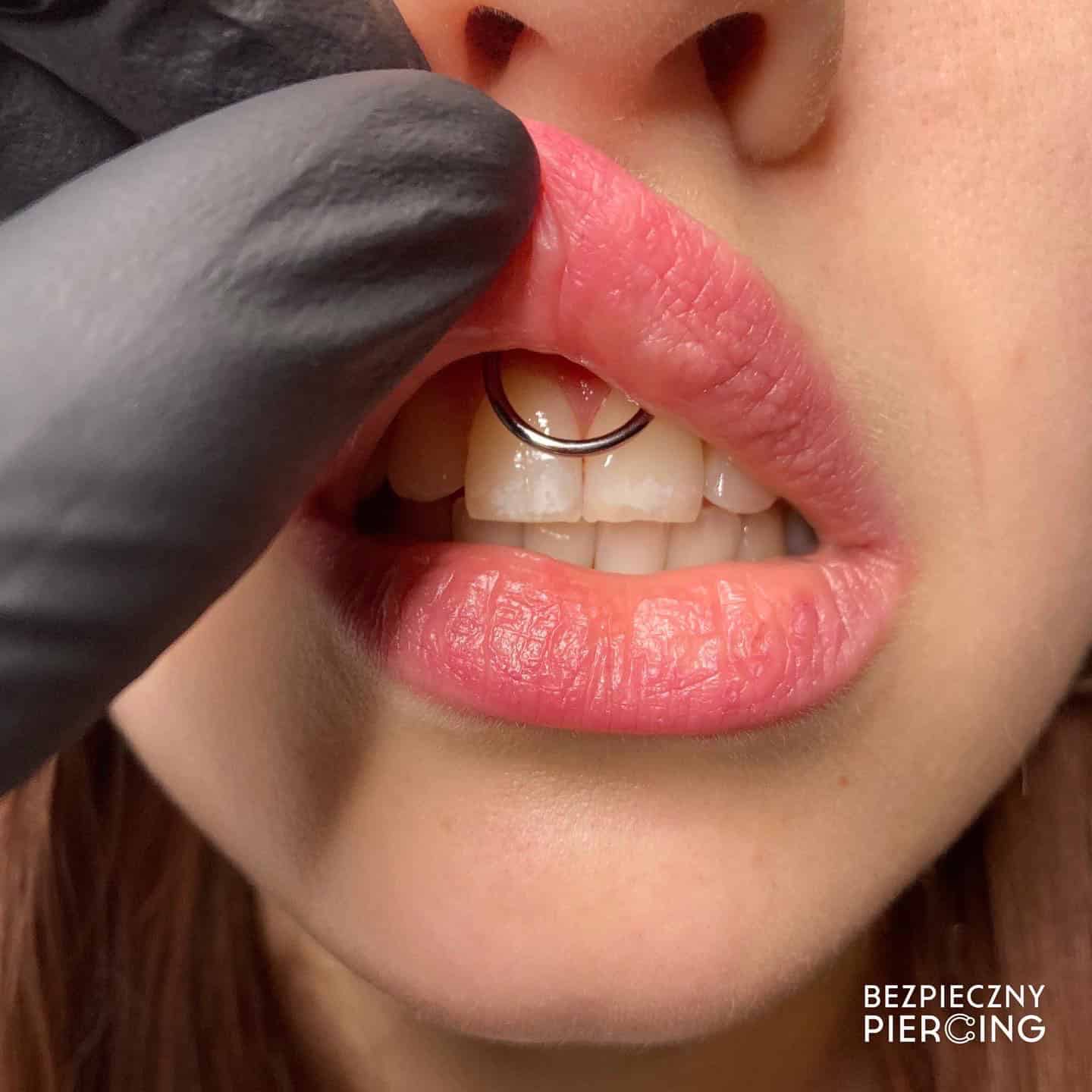 7sinstattoo.live Zbliżenie ust osoby pokazuje błyszczący kolczyk w kształcie pierścienia na wędzidełku pod górną wargą. Delikatnie unosi górną wargę ręką w rękawiczce, aby odsłonić piercing. Na zdjęciu w prawym dolnym rogu widnieje tekst „BEZPIECZNY PIERCING”, reprezentujący studio tatuaży Poznań i podkreślający produkt E9990DF4-F08C-4705-8F0B-A5C0146618FB. Najlepsze studio tatuażu Seven Sins Tattoo