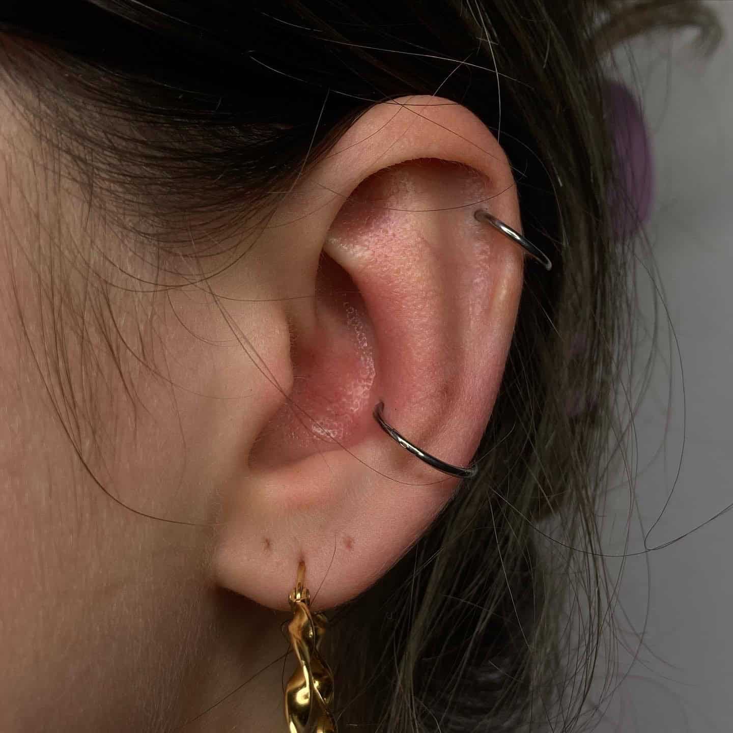 7sinstattoo.live Zbliżenie ucha osoby z podwójnym przekłuciem helisy ozdobionym dwoma prostymi srebrnymi kółkami, uzupełnione przekłuciem płatka ucha ze złotym skręconym kolczykiem kółkiem. Włosy są schludnie schowane za uchem, odsłaniając jasną skórę. Odkryj podobne style i bardziej kreatywne przekłucia w Studio Tatuażu Poznań, gdzie znajdziesz produkty takie jak F1AF7148-6E9F-4C2F-98B5-419D6172282E. Najlepsze studio tatuażu Seven Sins Tattoo