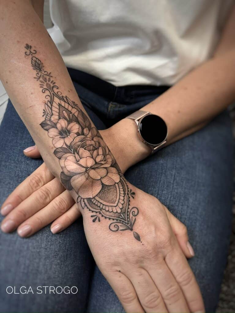 7sinstattoo.live Przedramię prezentujące IMG-5630, misterny czarno-szary kwiatowy tatuaż mandali rozciągający się na dłoń i nadgarstek. Osoba jest ubrana w biały top, niebieskie dżinsy i srebrny smartwatch. Ten misterny tatuaż, wykonany w Najlepszym Studio Tatuażu Poznań, zawiera szczegółowe kwiaty i ozdobne wzory z tekstem „OLGA STROGO. Najlepsze studio tatuażu Seven Sins Tattoo