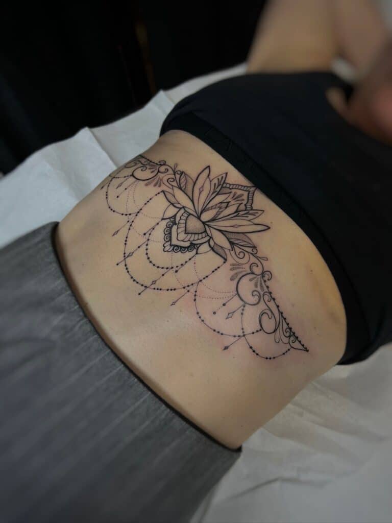 7sinstattoo.live W IMG-5736 szczegółowe zbliżenie ukazuje dolną część pleców osoby ozdobioną misternym tatuażem z cenionego studia tatuażu Poznań. Tatuaż przedstawia widoczny kwiat lotosu w centrum, otoczony misternymi, symetrycznymi wzorami i precyzyjnymi kropkami, które rozciągają się na dolnej części pleców. Osoba jest umieszczona na białym prześcieradle. Najlepsze studio tatuażu Seven Sins Tattoo