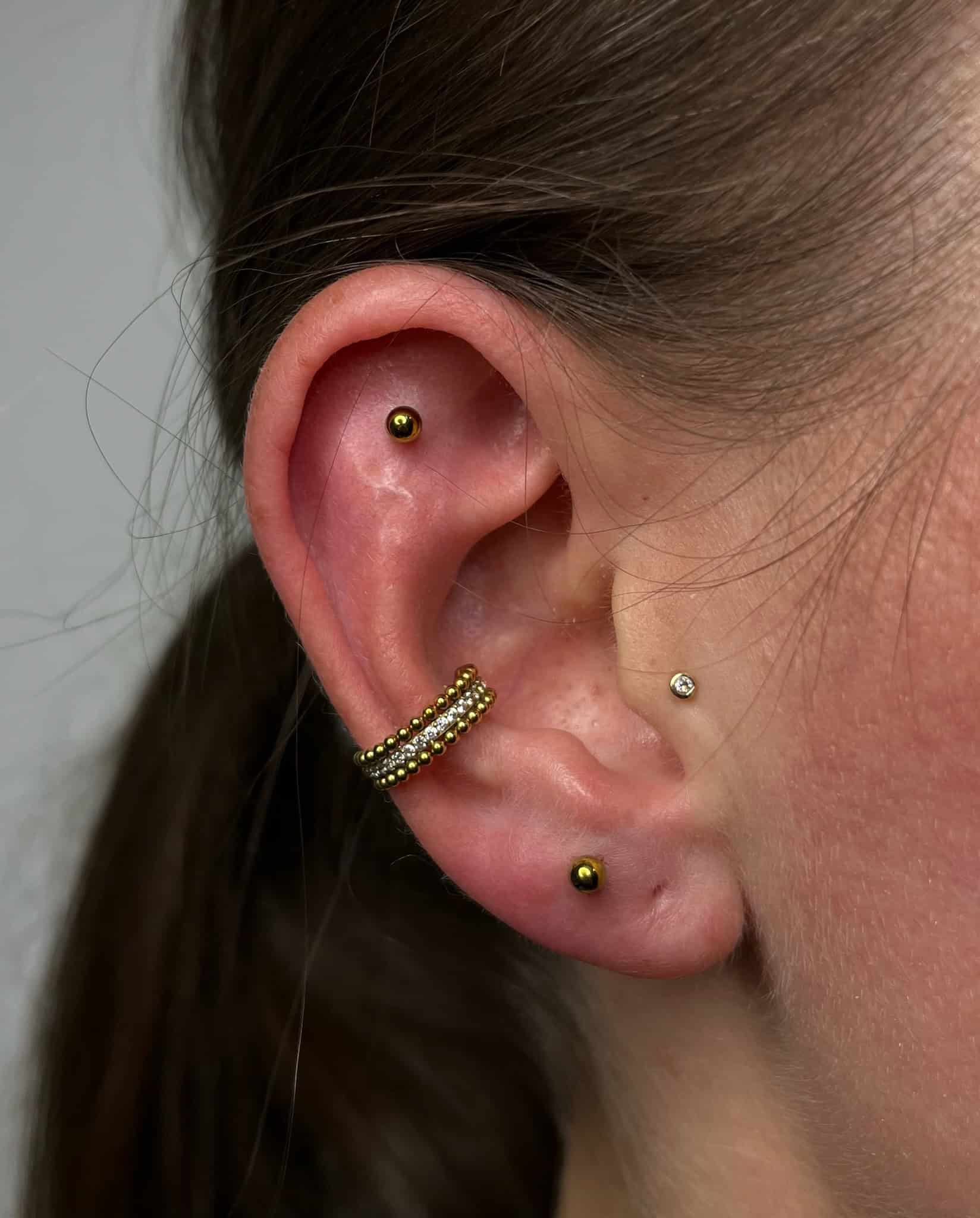 7sinstattoo.live Zbliżenie ucha osoby ozdobionego wieloma piercingami, z kółkiem na chrząstce w kolorze złotym, małym złotym kolczykiem w górnym uchu, małym diamentowym kolczykiem w płatku ucha i kolejnym złotym kolczykiem w dolnej chrząstce. Włosy osoby są związane z tyłu. Odkryj ten styl z IMG_0062 w najlepszym studiu tatuażu w Poznaniu. Najlepsze studio tatuażu Seven Sins Tattoo
