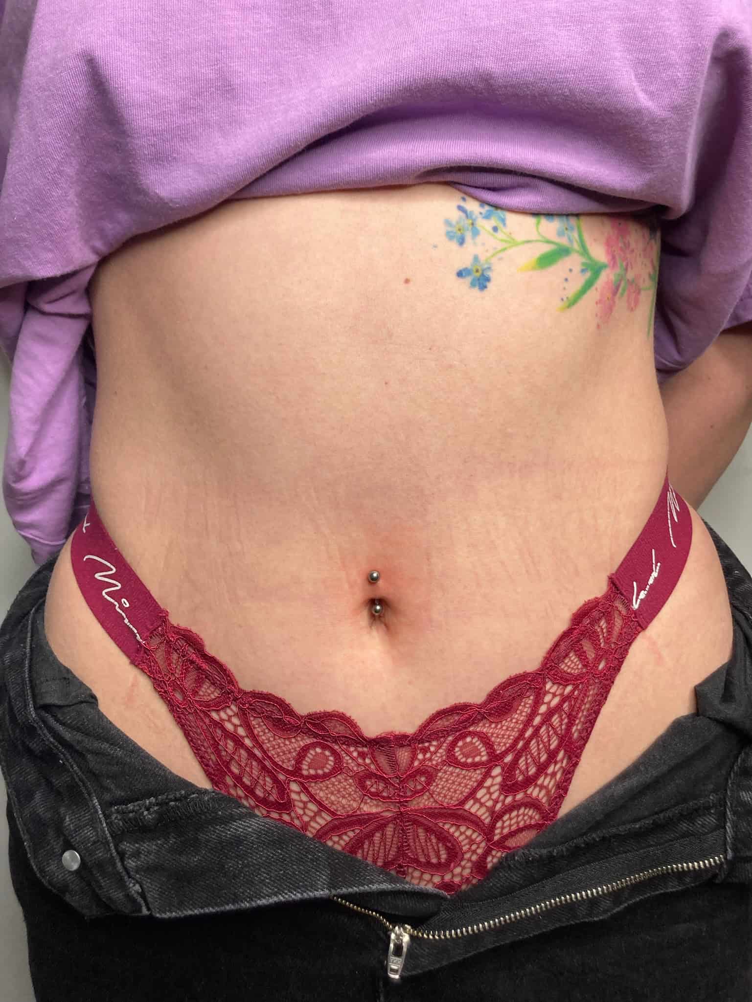 7sinstattoo.live Zbliżenie na dolną część brzucha i talię osoby ukazuje czerwone koronkowe stringi wystające z rozpiętych czarnych dżinsów. Osoba ta ma piercing w pępku i może pochwalić się żywym kwiatowym tatuażem ze Studio Tatuażu Poznań na boku. Ma na sobie lekko uniesioną fioletową koszulę, uchwyconą na zdjęciu o nazwie IMG_0189. Najlepsze studio tatuażu Seven Sins Tattoo