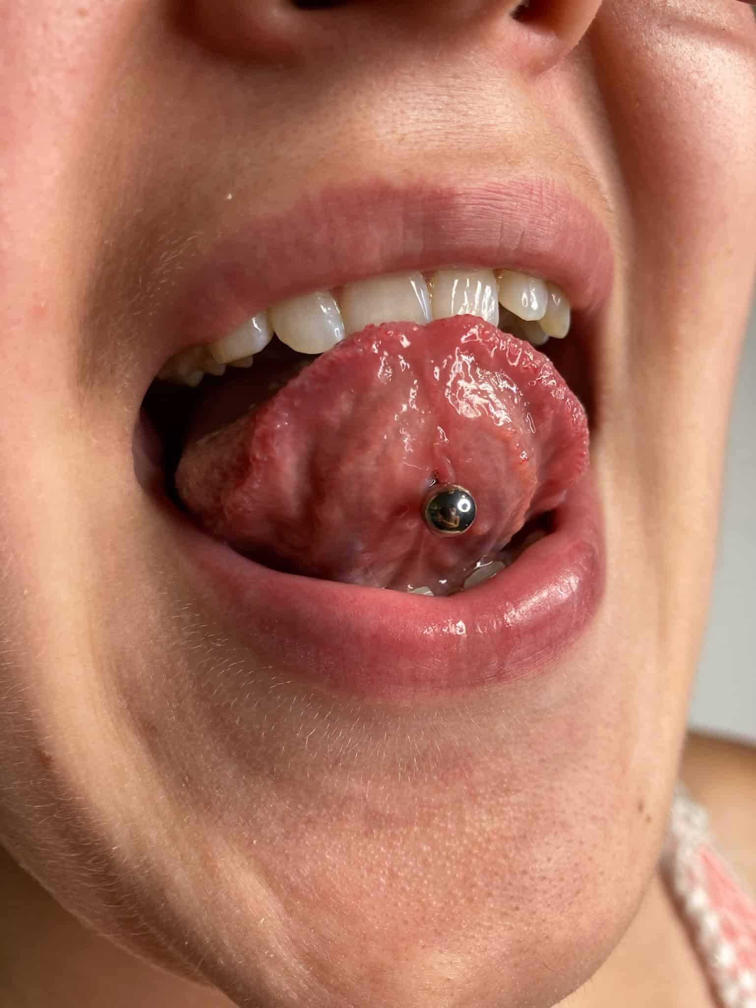 7sinstattoo.live Zbliżenie osoby wystawiającej język, pokazujące srebrny kolczyk do języka. Wilgotny język jest podkreślony lekko rozchylonymi wargami i widocznymi zębami, podczas gdy gładka skóra wokół ust sugeruje staranną pielęgnację, przypominając jakość, jakiej można się spodziewać po IMG_0368. Najlepsze studio tatuażu Seven Sins Tattoo