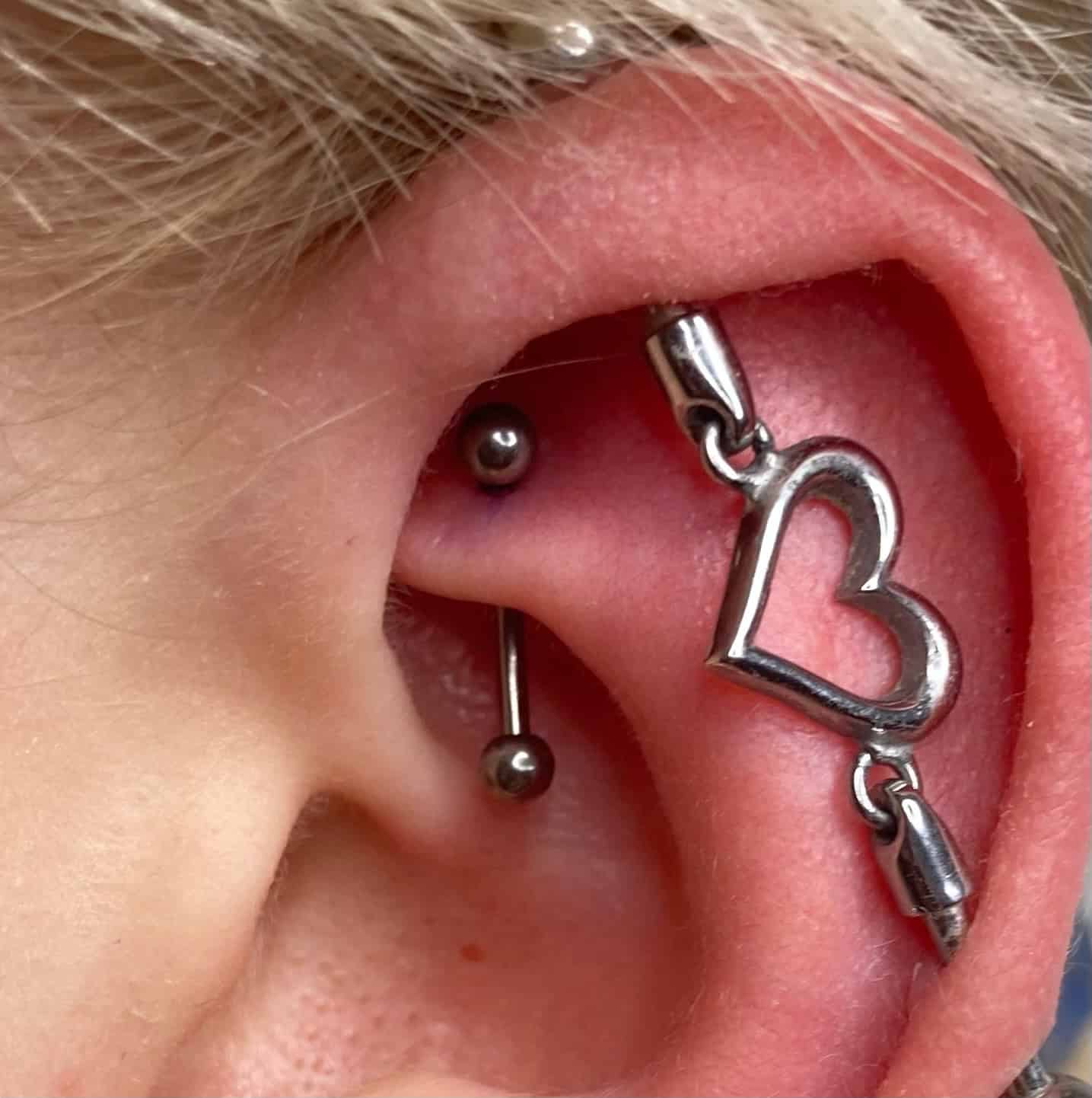 7sinstattoo.live Zbliżenie ukazuje ucho osoby ozdobione wieloma piercingami, w tym industrialnym piercingiem z metalowym prętem. Dodatkowo, zwisający kolczyk z zawieszką w kształcie serca zwisa z górnej części ucha. Blond włosy osoby podkreślają odważny styl, idealny do zaprezentowania w najlepszym studio tatuażu w Poznaniu. Ta scena doskonale oddaje IMG_0646. Najlepsze studio tatuażu Seven Sins Tattoo