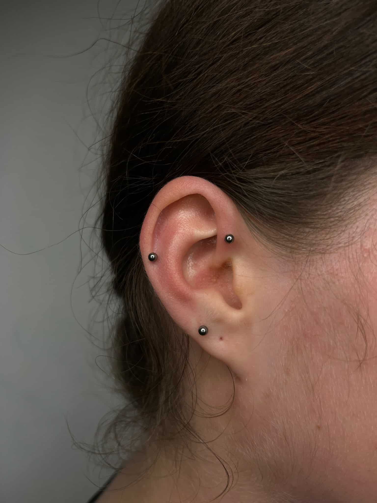 7sinstattoo.live Zbliżenie ucha osoby z dwoma małymi metalowymi kolczykami w kształcie kulek: jednym na górnej chrząstce i jednym na płatku ucha. Włosy są związane z tyłu, wyraźnie odsłaniając ucho na neutralnym tle w IMG_0741, najlepsze studio tatuażu Poznań. Najlepsze studio tatuażu Seven Sins Tattoo