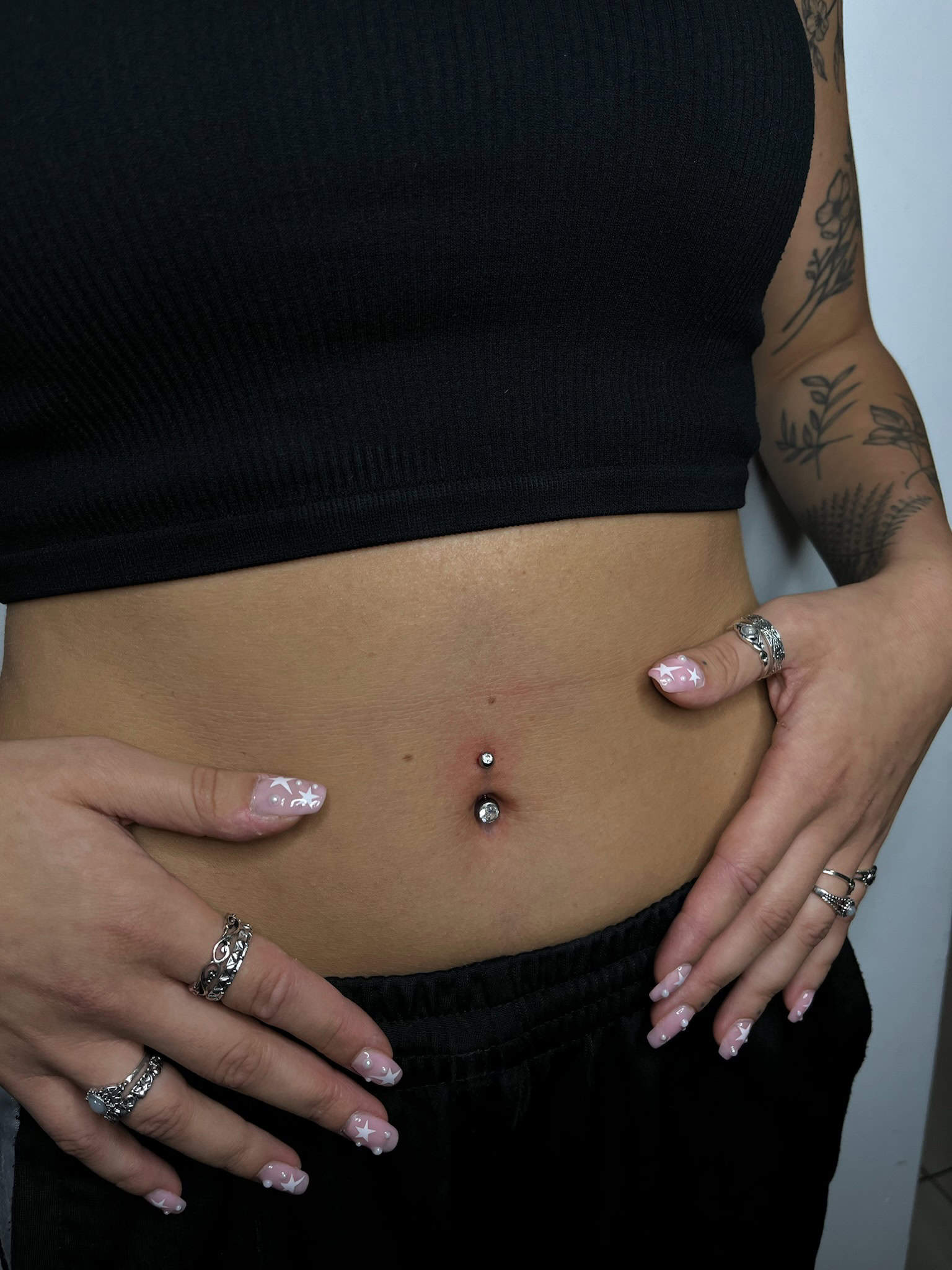 7sinstattoo.live Osoba pewnie eksponuje piercing pępka ozdobiony dwoma srebrnymi ćwiekami, mając na sobie czarny krótki top i czarne spodnie. Dłonie tej osoby, ozdobione pierścionkami i zadbane paznokcie z jasnoróżowym lakierem i kwiatowymi wzorami, odsłaniają widoczne tatuaże na przedramieniu ze studia tatuażu Poznań. Ten uderzający obraz został uchwycony w produkcie IMG_0836. Najlepsze studio tatuażu Seven Sins Tattoo