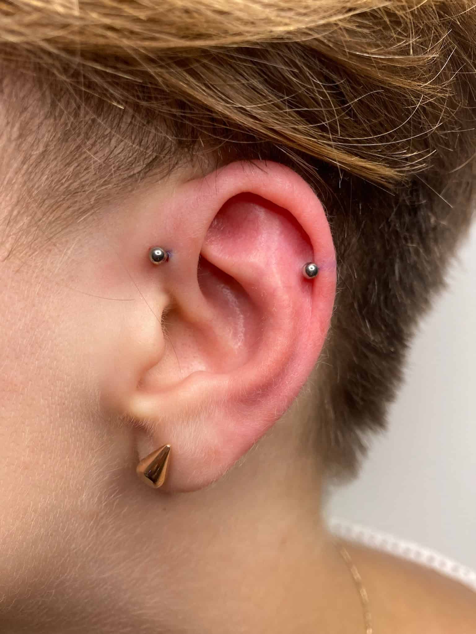 7sinstattoo.live Zbliżenie ucha osoby z dwoma przekłuciami wewnętrznej chrząstki i przekłuciem helix, wszystkie z małymi, srebrnymi metalowymi ćwiekami (IMG_0837). Osoba ma również złoty, trójkątny kolczyk w płatku ucha. Z krótkimi, jasnobrązowymi włosami prezentuje swój styl inspirowany klimatem studia tatuaży Poznań. Najlepsze studio tatuażu Seven Sins Tattoo