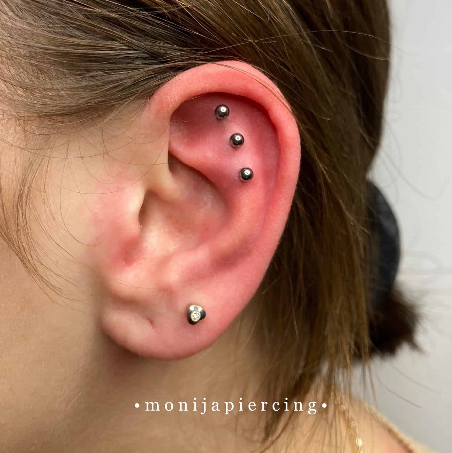 7sinstattoo.live Zbliżenie ucha z czterema kolczykami. Trzy małe kolczyki kulkowe są ustawione pionowo wzdłuż górnej chrząstki, a pojedynczy kolczyk z przezroczystym klejnotem zdobi płatek ucha. Włosy są elegancko odgarnięte do tyłu, podkreślając kolczyki w miejscu, które można zidentyfikować jako najlepsze studio tatuażu Poznań. Tło pozostaje neutralne. Tekst: "@monijapiercing 
Nazwa produktu: IMG_0859. Najlepsze studio tatuażu Seven Sins Tattoo