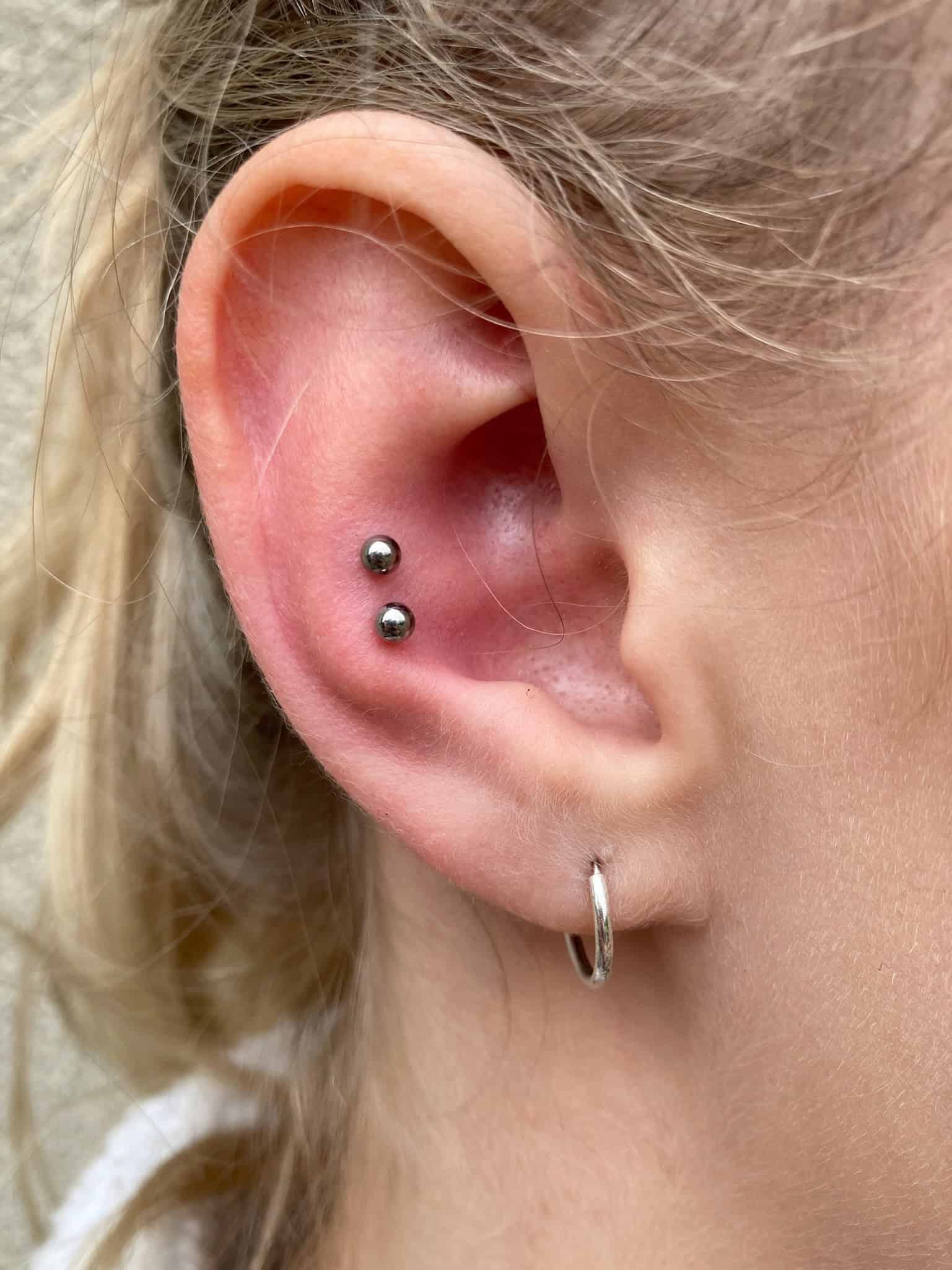 7sinstattoo.live Zdjęcie z bliska zatytułowane IMG_1041 (1), przedstawiające ucho ozdobione dwoma srebrnymi kolczykami na górnej chrząstce i srebrnym kolczykiem-kółkiem na płatku ucha. Osoba ta ma jasnobrązowe włosy ułożone luźno na neutralnej beżowej ścianie, prawdopodobnie sfotografowane w studio tatuażu Poznań. Najlepsze studio tatuażu Seven Sins Tattoo