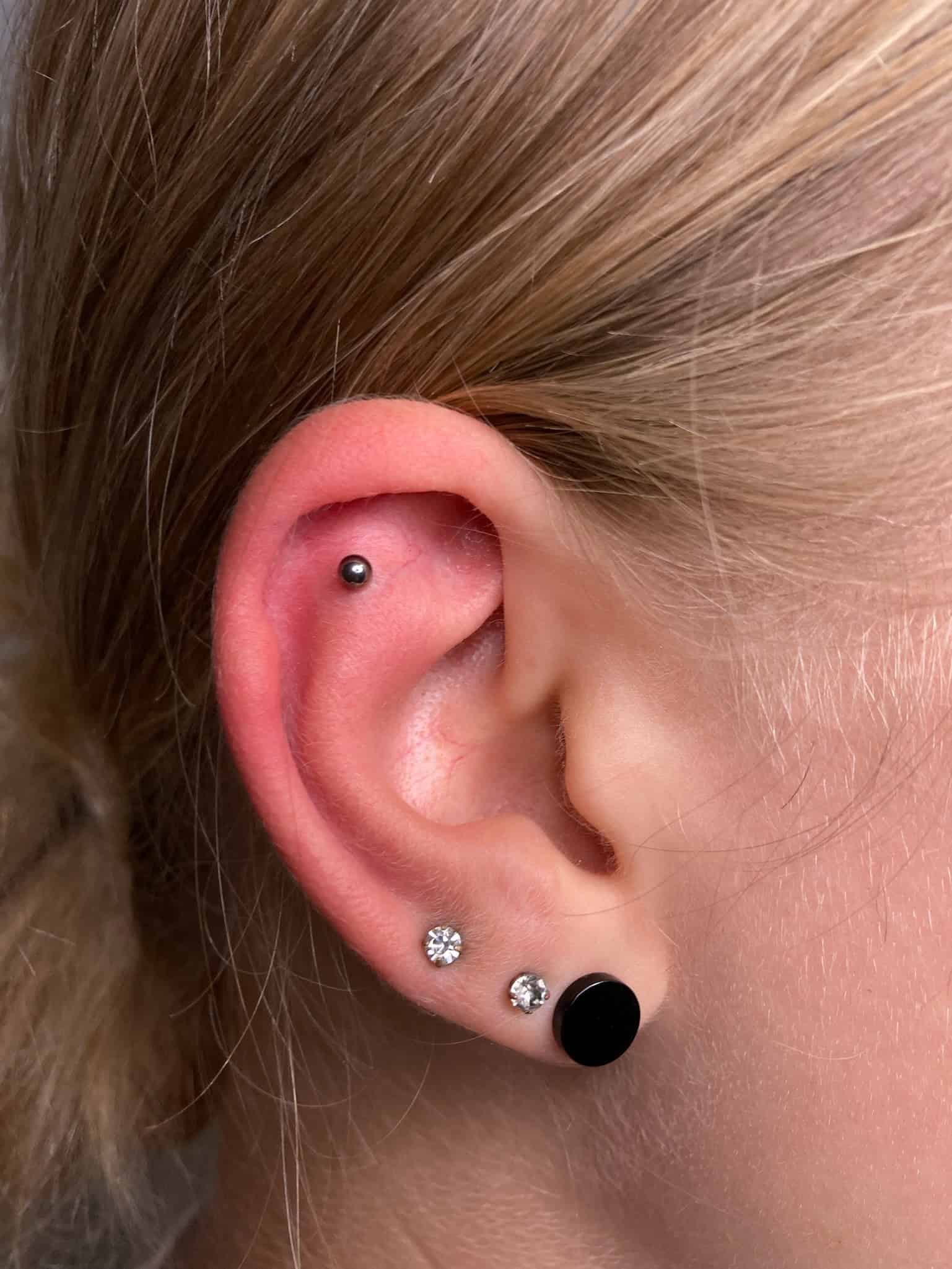 7sinstattoo.live Zbliżenie ucha osoby z wieloma piercingami ze studia tatuażu Poznań, z Product IMG_1418. Ucho prezentuje mały srebrny kolczyk na górnej chrząstce, kolczyk z diamentem na środkowym płacie i czarny okrągły kolczyk na dolnym płacie. Tło jest rozmyte. Osoba ma jasne włosy. Najlepsze studio tatuażu Seven Sins Tattoo