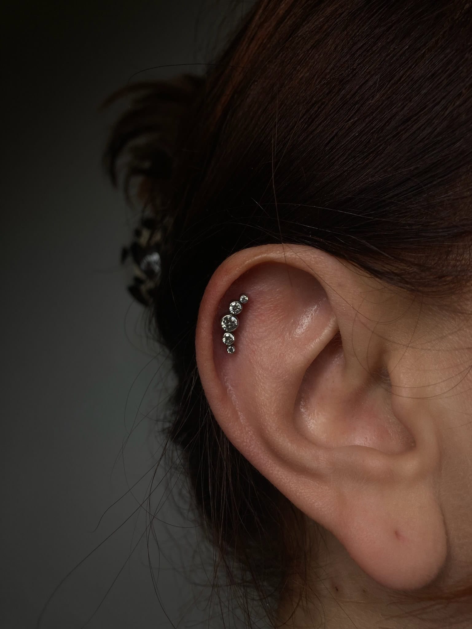 7sinstattoo.live Zbliżenie na ucho osoby z przekłuciem chrząstki helix i rzędem pięciu małych, błyszczących kolczyków. Luźne pasma brązowych włosów oprawiają ucho, a tło stanowi ciemny, rozmyty gradient — mistrzowsko uchwycony w studio tatuażu Poznań w IMG_1546. Najlepsze studio tatuażu Seven Sins Tattoo
