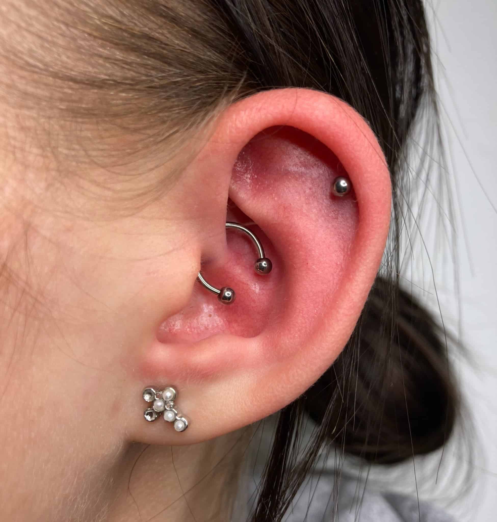 7sinstattoo.live Na pierwszym planie widoczne jest ucho osoby ozdobione wieloma piercingami, z zakrzywionym sztangą w górnej chrząstce i małym kolczykiem w wewnętrznej muszli. Płatek ucha prezentuje elegancki kolczyk w kształcie kwiatu z płatkami pereł. Neutralne tło sugeruje, że zdjęcie zostało zrobione w najlepszym poznańskim studiu piercingu i salonie tatuażu pod nazwą produktu IMG_1633. Najlepsze studio tatuażu Seven Sins Tattoo
