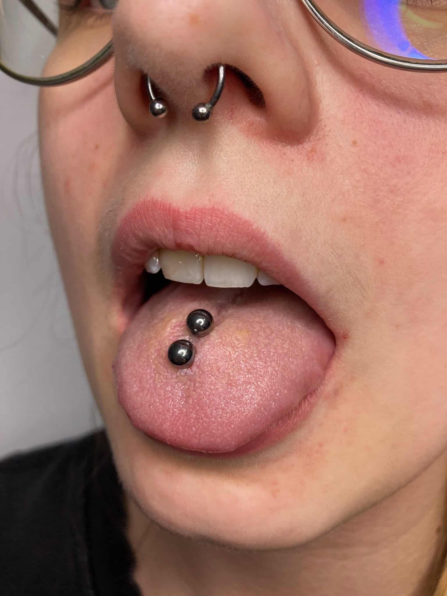 7sinstattoo.live Zbliżenie osoby wystawiającej język, pokazującej dwa kolczyki z czarną biżuterią z kulkami. Ma również piercing przegrody nosowej z dwoma srebrnymi kulkami i nosi okulary. Przy jasnej karnacji, to może być uchwycone w najlepszym studio tatuażu w Poznaniu, obraz o nazwie „IMG_1754. Najlepsze studio tatuażu Seven Sins Tattoo