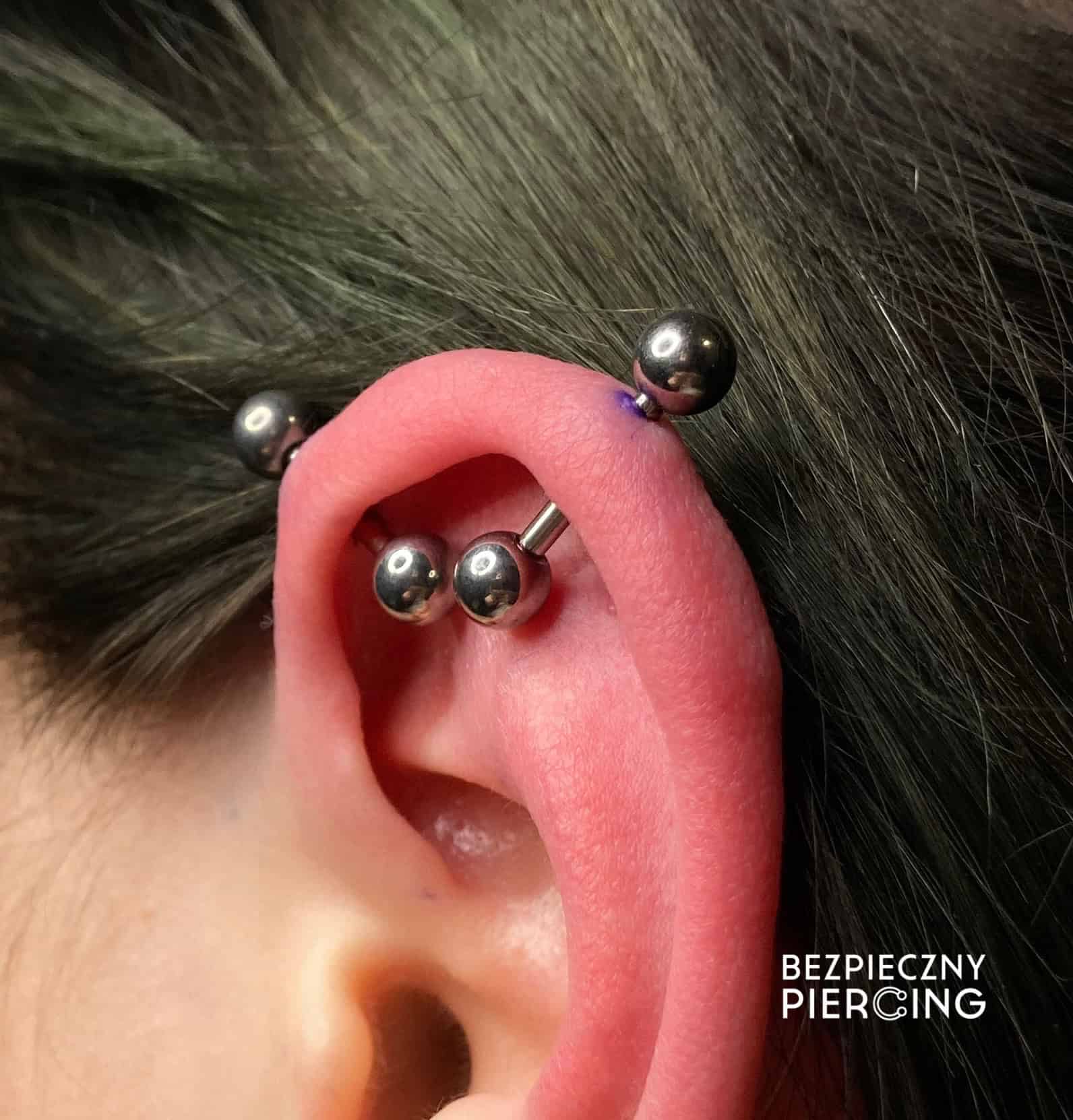 7sinstattoo.live Zbliżenie ucha ozdobionego wieloma piercingami zaprezentowanymi w IMG_2003. Ucho ma trzy ułożone kolczyka barbell wzdłuż zewnętrznej krawędzi, co świadczy o kunszcie studia tatuażu Poznań. Piercingi wydają się być wykonane profesjonalnie. W prawym dolnym rogu napisano białym tekstem „BEZPIECZNY PIERCING”. Najlepsze studio tatuażu Seven Sins Tattoo