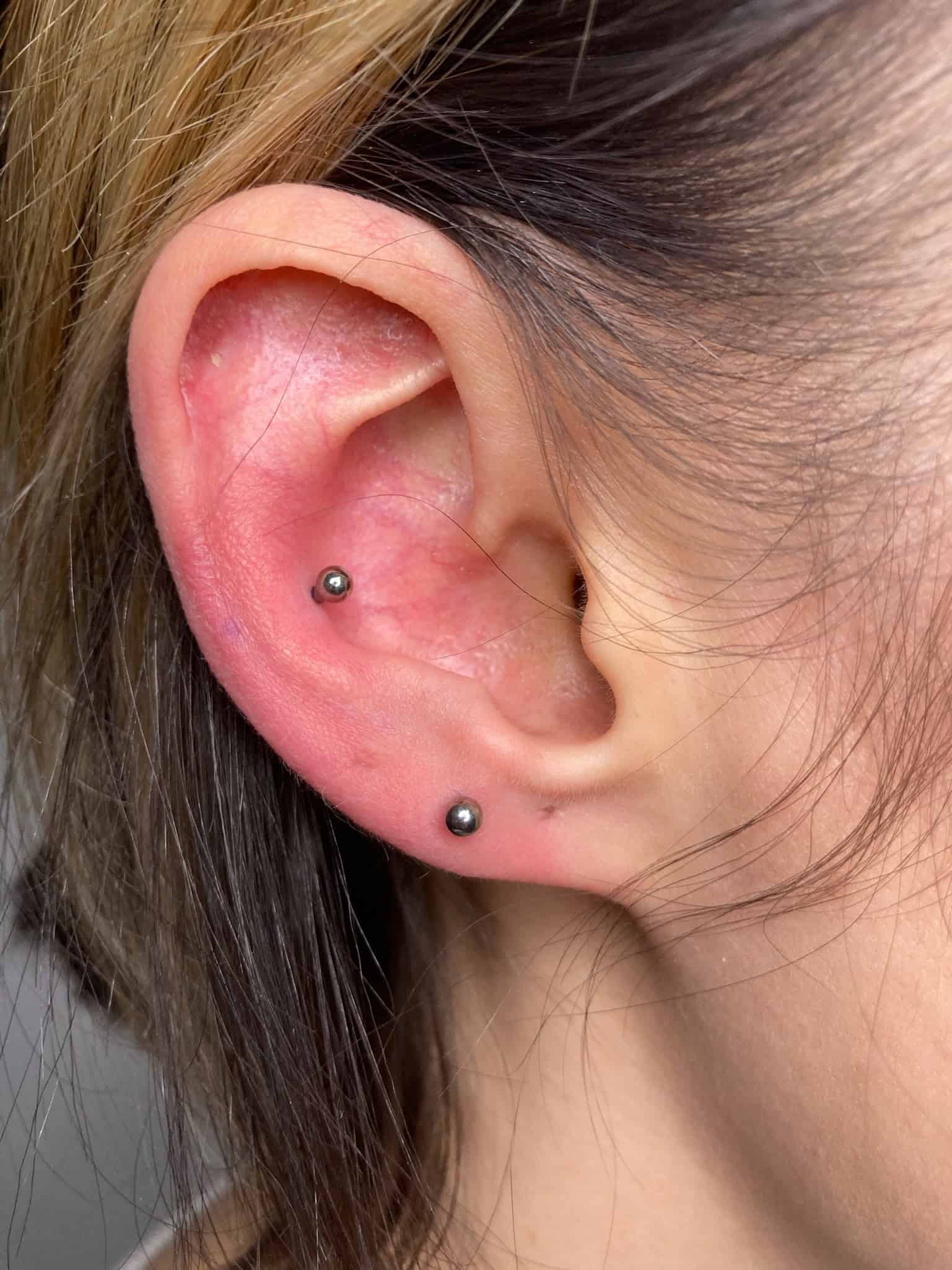 7sinstattoo.live Zbliżenie ucha osoby z dwoma piercingami: jednym na górnej wewnętrznej chrząstce i jednym na dolnym płatku ucha, każdy z małymi, ciemnymi metalowymi ćwiekami. Jasnobrązowe włosy osoby są częściowo widoczne w ramce. Odkryj wyjątkowe style w studio tatuaży Poznań, aby uzyskać kompletny wygląd z IMG_2107! Najlepsze studio tatuażu Seven Sins Tattoo