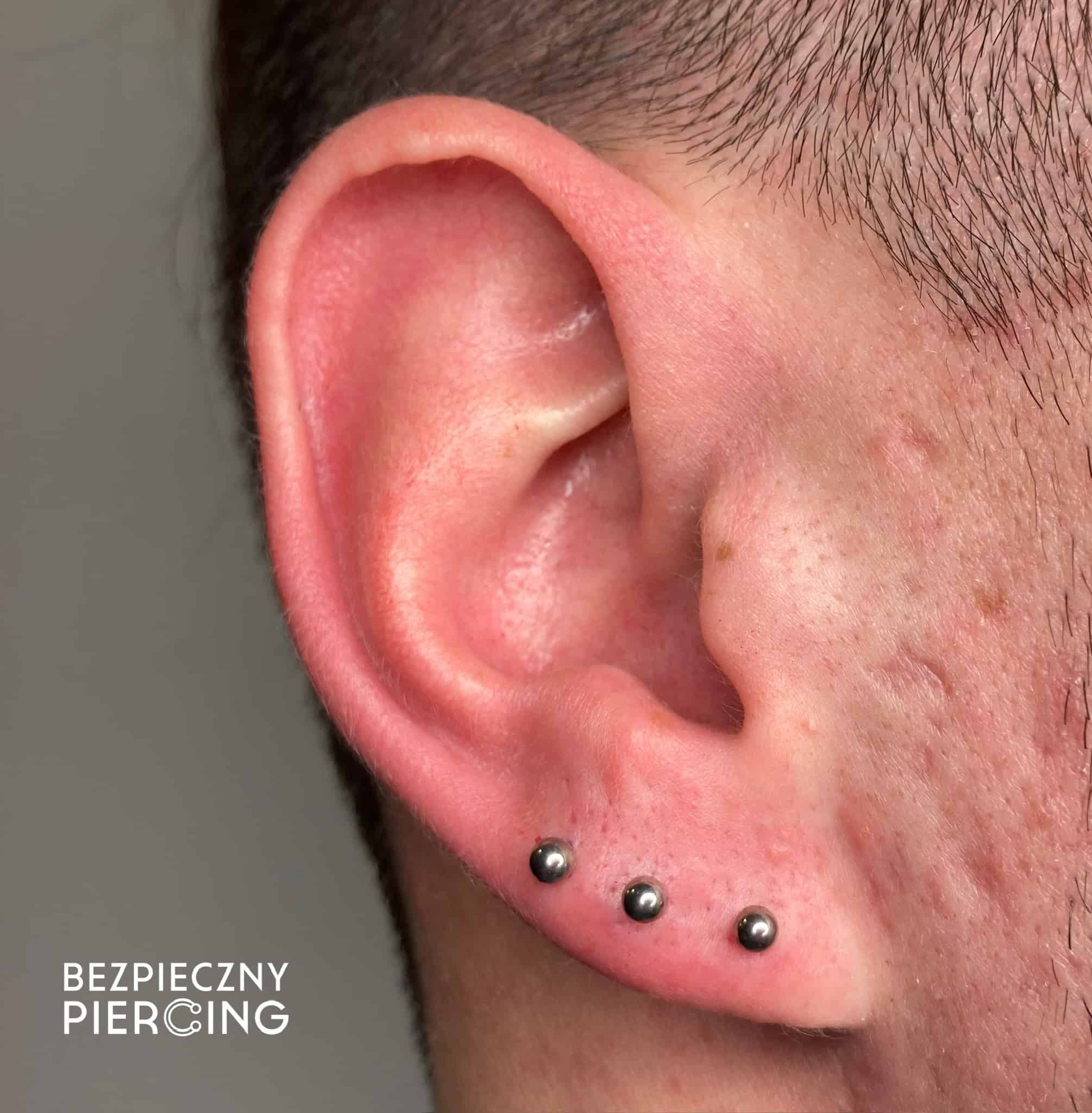 7sinstattoo.live Zbliżenie ucha osoby z trzema srebrnymi kulkami w płatku. Tło jest neutralne, a w lewym dolnym rogu widoczny jest tekst „BEZPIECZNY PIERCING”. Skóra w pobliżu piercingu jest lekko różowa, co sugeruje niedawne przekłucie w studio tatuaży Poznań. Nazwa produktu: IMG_2232. Najlepsze studio tatuażu Seven Sins Tattoo