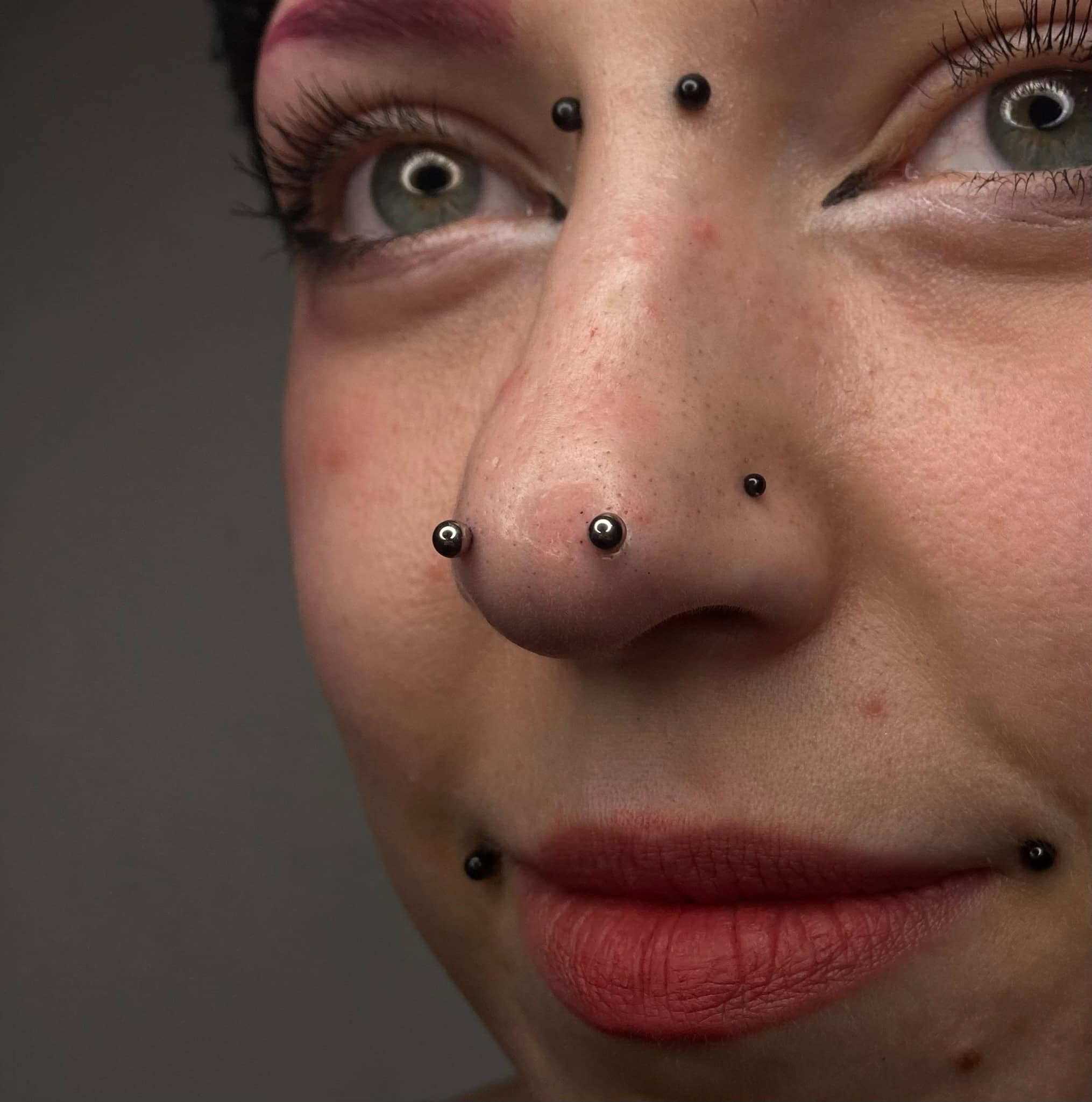 7sinstattoo.live Zbliżenie twarzy osoby z wieloma piercingami twarzy, w tym dwoma kolczykami w nosie, kolczykiem w przegrodzie nosowej i parą piercingów wargi dolnej. Osoba ma różowy makijaż oczu, skrzydlatą kreskę na powiece i jasnoczerwoną szminkę. Odkryj ten styl na naszym wyróżnionym zdjęciu: IMG_2240 w najlepsze studio tatuażu Poznań. Najlepsze studio tatuażu Seven Sins Tattoo