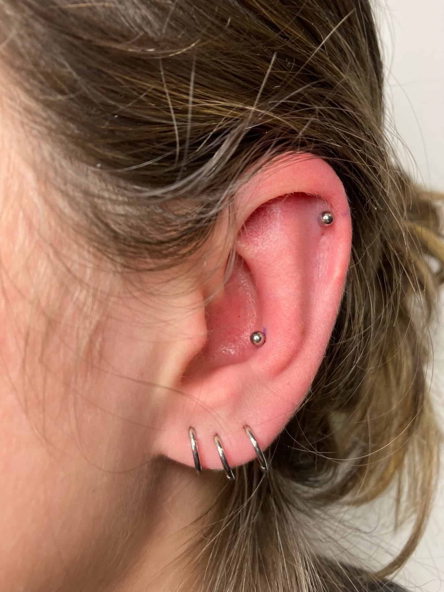 7sinstattoo.live Zbliżenie ucha osoby z trzema piercingami stylizowanymi za pomocą IMG_2278. Dwa małe kolczyki w kształcie kółek zdobią dolny płatek ucha, a pojedynczy kolczyk zdobi helisę (górną chrząstkę ucha). Jasnobrązowe włosy osoby są częściowo odgarnięte do tyłu, co zapewnia wypolerowany wygląd przypominający kunszt studio tatuaży Poznań. Najlepsze studio tatuażu Seven Sins Tattoo