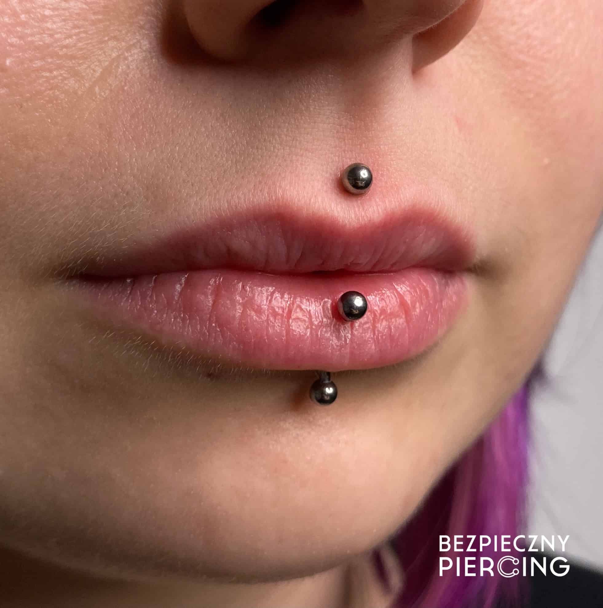 7sinstattoo.live Zbliżenie na zdjęcie zatytułowane IMG_2488 przedstawia usta osoby z trzema piercingami: jednym na rynience nosowej nad górną wargą i dwoma na dolnej wardze. Osoba ma naturalny odcień skóry i różowe usta, z widocznymi fioletowymi włoskami w prawym dolnym rogu. Na zdjęciu widoczny jest tekst „BEZPIECZNY PIERCING” z Najlepszego Studia Tatuażu Poznań. Najlepsze studio tatuażu Seven Sins Tattoo