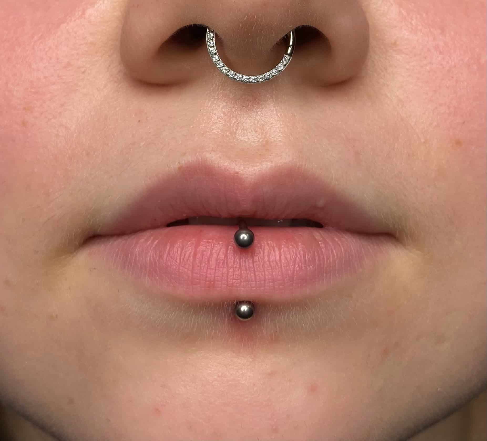 7sinstattoo.live Zbliżenie dolnej części twarzy osoby prezentujące IMG_2930, z piercingiem przegrody nosowej ozdobionym okrągłym, wysadzanym klejnotami pierścieniem i pionowym piercingiem wargi dolnej z dwoma małymi, czarnymi kulkami. Gładka skóra i naturalny kolor ust podkreślają kunszt, podkreślając prace z najlepszego studia tatuaż w Poznaniu. Najlepsze studio tatuażu Seven Sins Tattoo