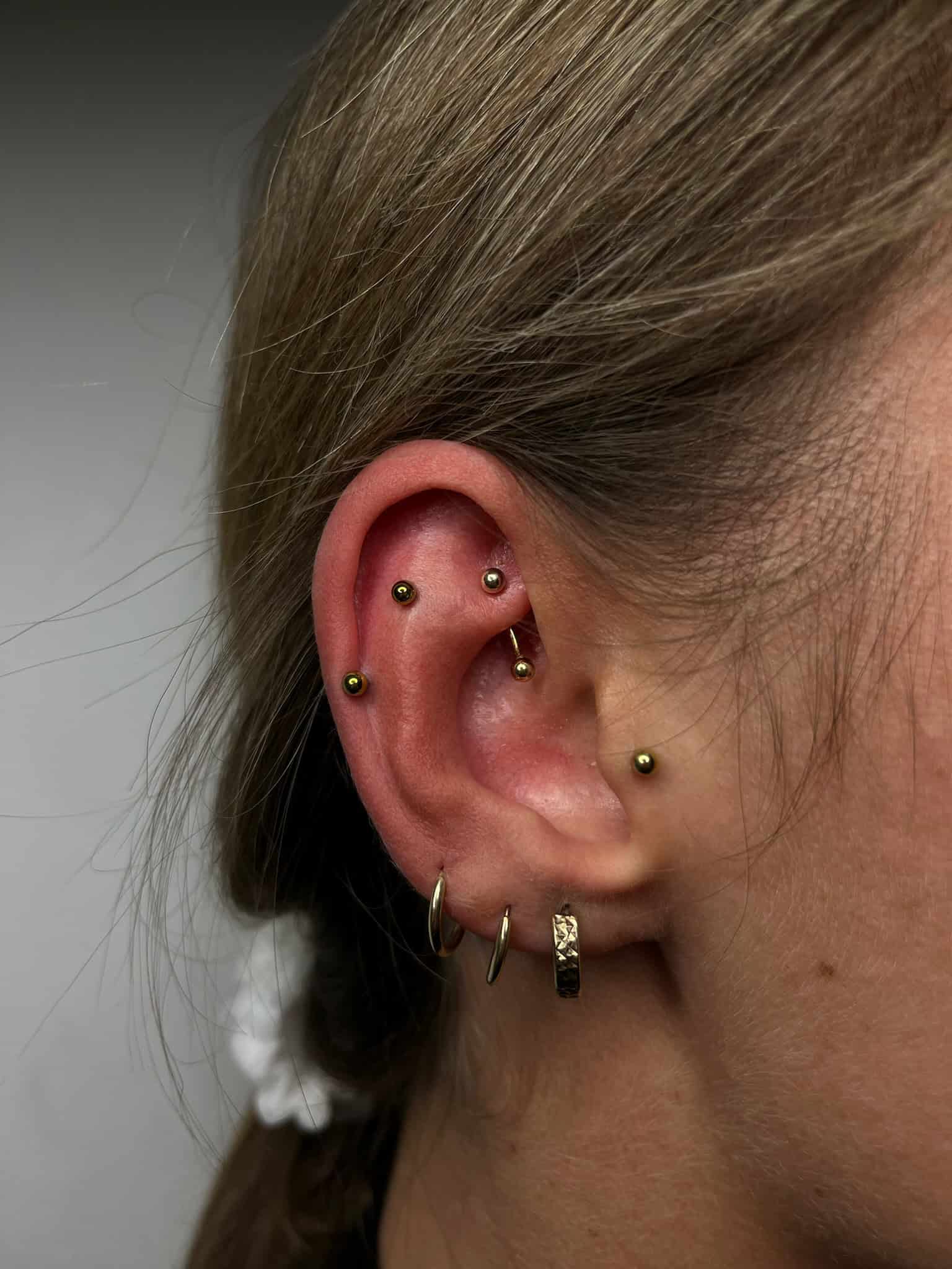 7sinstattoo.live Zbliżenie z IMG_3037 pokazuje ucho ozdobione wieloma piercingami, idealne dla osób odwiedzających najlepsze studio tatuażu w Poznaniu. Ucho ma kilka złotych kolczyków wzdłuż chrząstki i kilka złotych kółek na płatku ucha. Włosy osoby są ściągnięte do tyłu, zaakcentowane białym ozdobą do włosów. Najlepsze studio tatuażu Seven Sins Tattoo