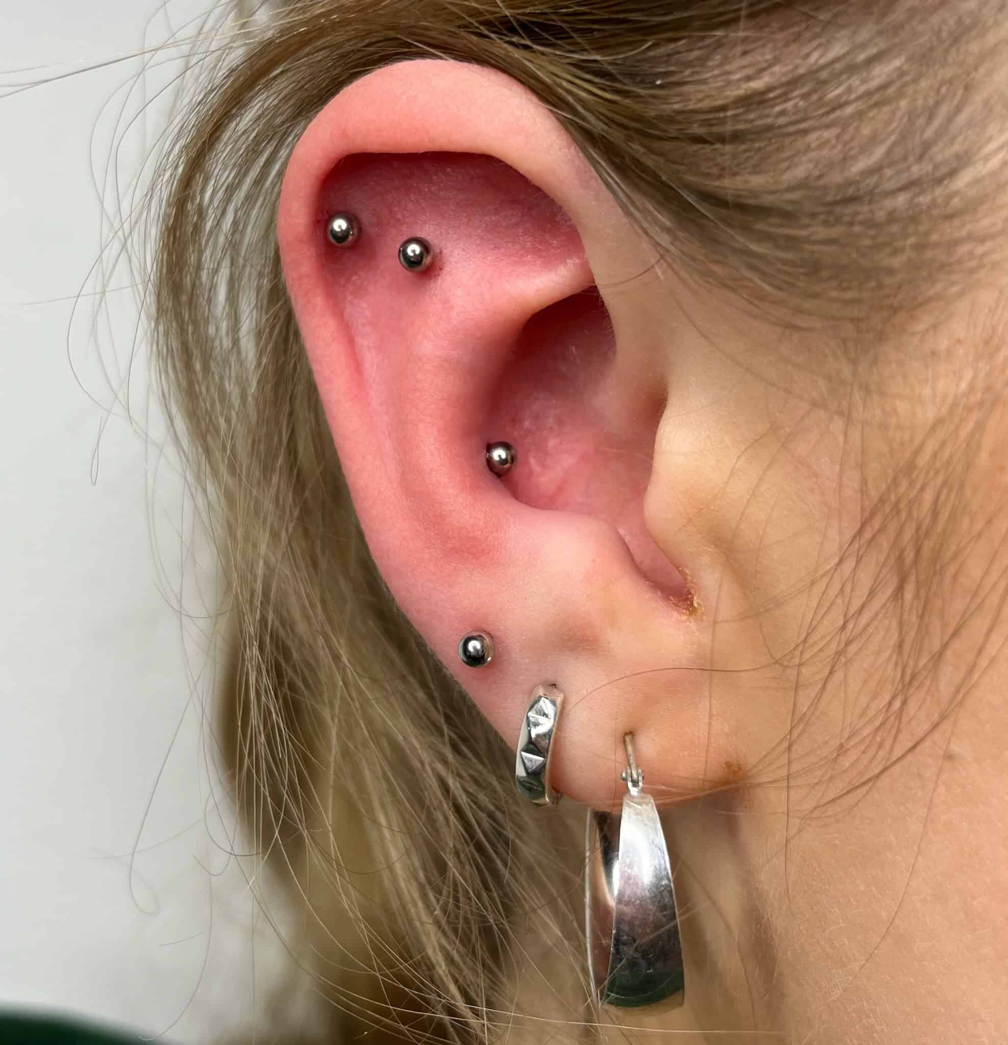 7sinstattoo.live Opis produktu dla IMG_3100: 
Zbliżenie lewego ucha osoby ozdobionego wieloma piercingami, zrobione w studio tatuażu Poznań. Ucho ma dwa małe, srebrne ćwieki na górnej części, mały kółko obok wewnętrznego ucha i dwa srebrne kolczyki w kształcie kółek na dolnym płatku — jeden gładki i jeden teksturowany. Najlepsze studio tatuażu Seven Sins Tattoo