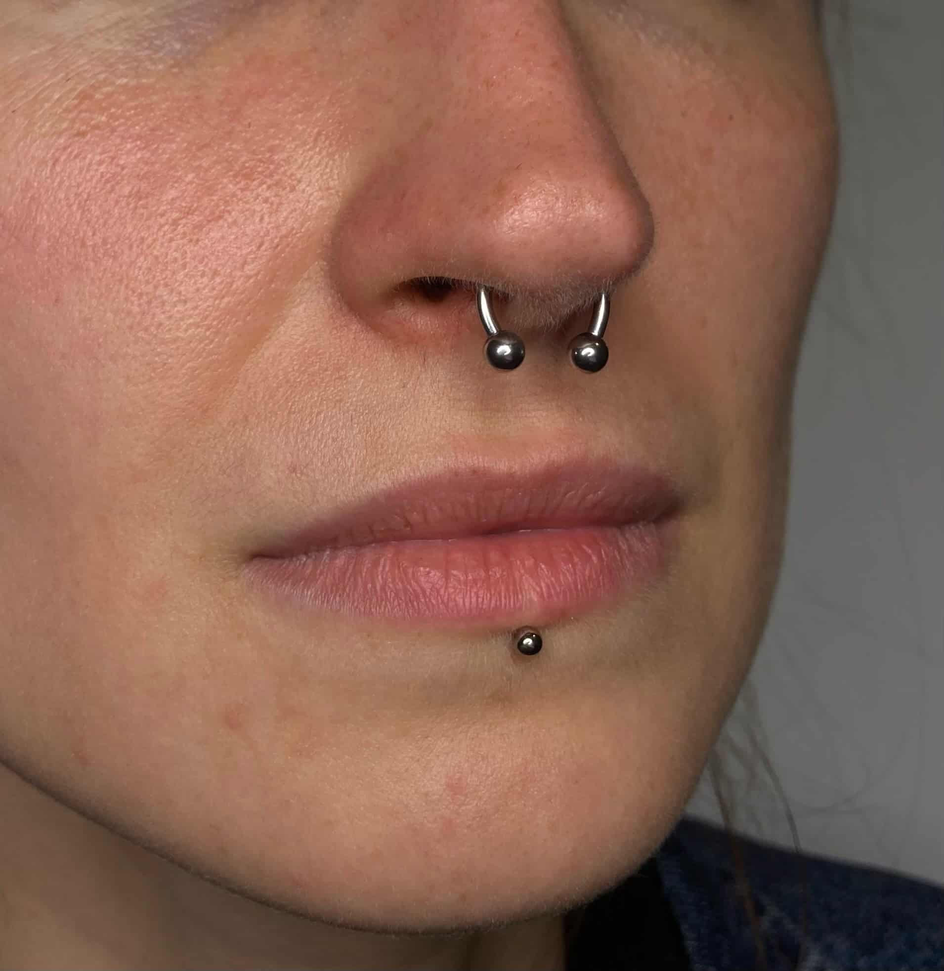 7sinstattoo.live Szczegółowe zdjęcie z bliska, IMG_3131, uchwyca twarz osoby, skupiając się na jej nosie i ustach. Osoba ta nosi przekłucie przegrody nosowej z okrągłym sztangą, uzupełnione o przekłucie wargi dolnej. Jej naturalna cera jest pięknie podkreślona widocznymi piegami i porami, artystycznie zaprezentowanymi w studio tatuażu Poznań. Najlepsze studio tatuażu Seven Sins Tattoo