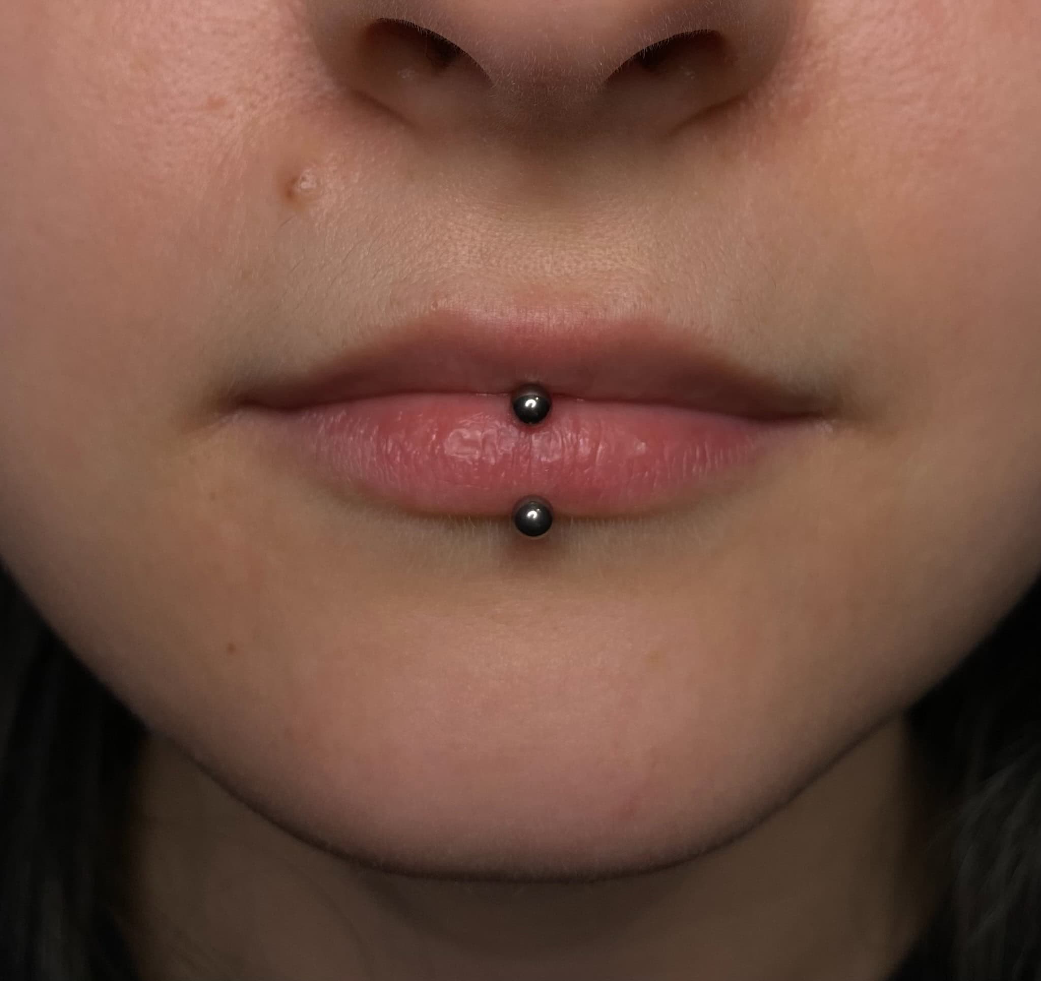 7sinstattoo.live Zbliżenie ust osoby z pionowym piercingiem wargi. Piercing składa się z dwóch małych, czarnych kulek — jednej na dolnej wardze, a drugiej poniżej — połączonych prętem. Uchwycony w IMG_3158 w najlepsze studio tatuażu Poznań, tło nie jest widoczne, skupiając się tylko na dolnej części twarzy. Najlepsze studio tatuażu Seven Sins Tattoo