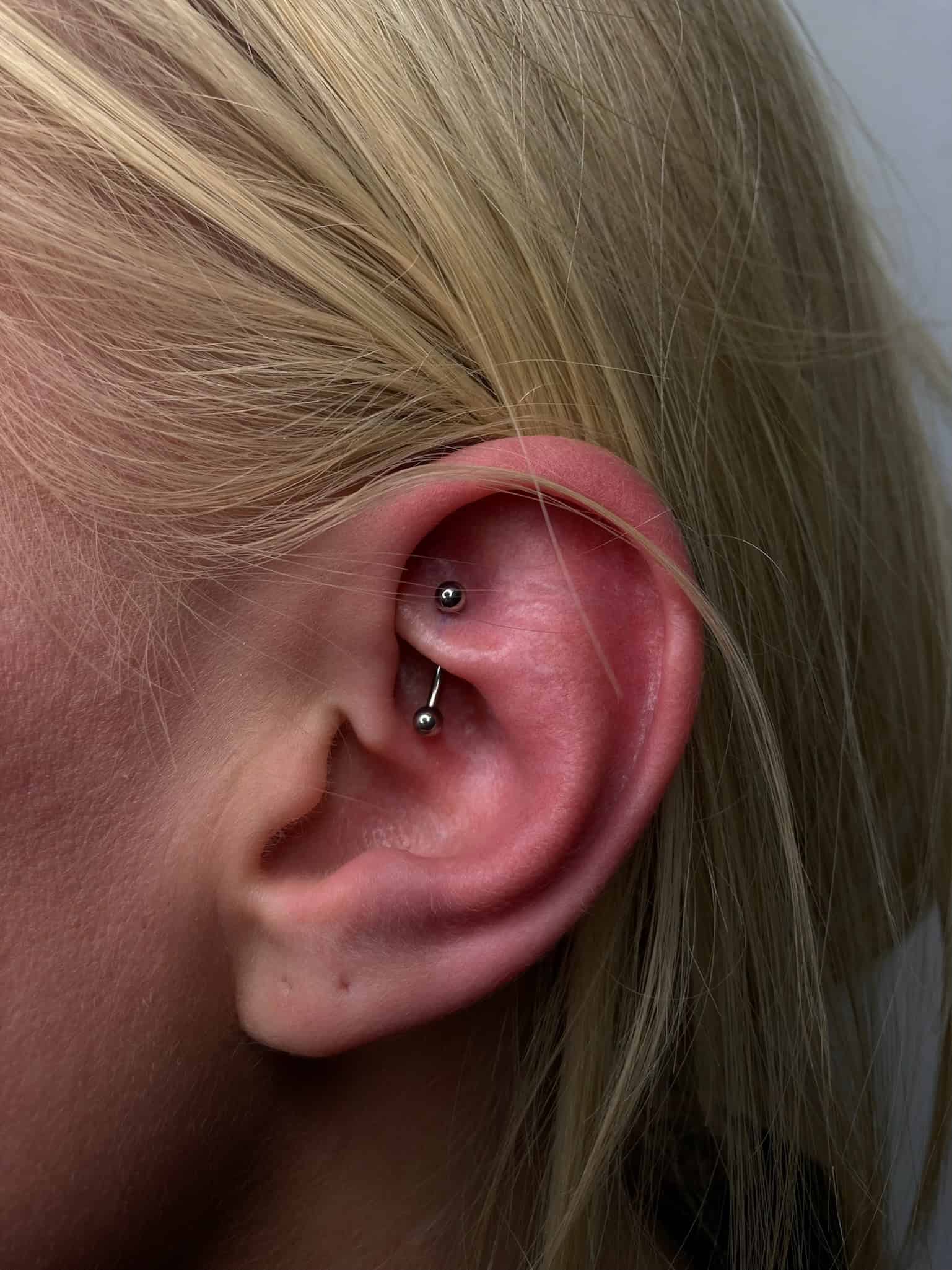 7sinstattoo.live Zbliżenie ucha ozdobionego świeżym industrialnym piercingiem ze studia tatuażu Poznań, z produktem IMG_3179. Piercing prezentuje prosty metalowy sztangę z dwoma widocznymi srebrnymi kulkami na każdym końcu. Ucho wydaje się lekko czerwone, co wskazuje na niedawny piercing, a osoba ma blond włosy. Najlepsze studio tatuażu Seven Sins Tattoo