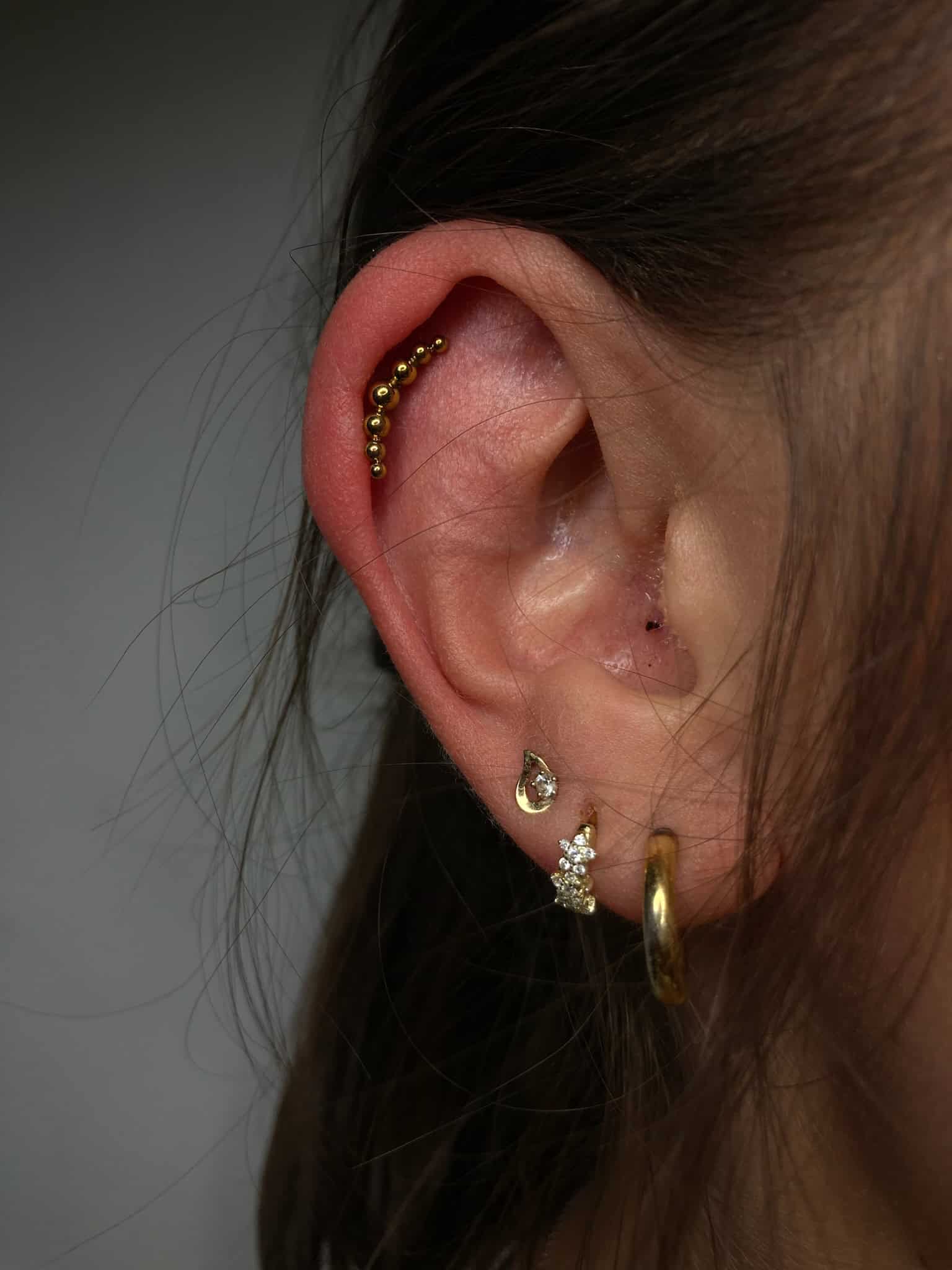 7sinstattoo.live Zbliżenie na ludzkie ucho ozdobione wieloma piercingami, prezentujące różne kolczyki, w tym zakrzywiony rząd małych złotych koralików wzdłuż helisy, gwiazdę, półksiężyc, mały sztyft i prosty złoty kółko na płatku uchwycony idealnie na zdjęciu IMG_3569 autorstwa studia tatuażu Poznań. Najlepsze studio tatuażu Seven Sins Tattoo