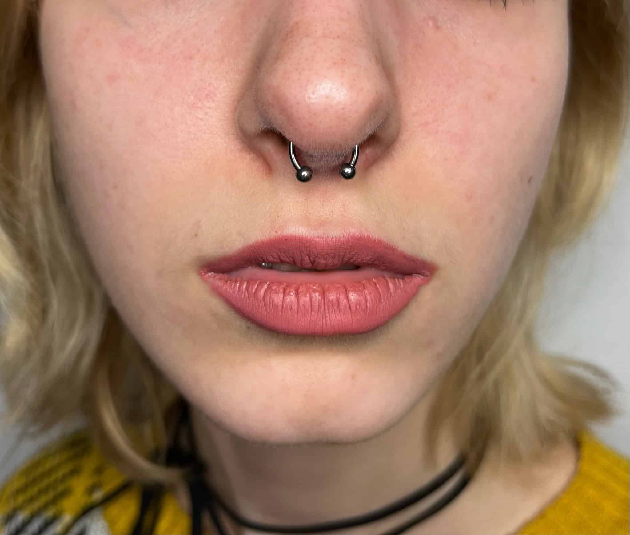 7sinstattoo.live Zbliżenie twarzy osoby, skupiające się na nosie i ustach. Osoba ma piercing przegrody nosowej z kółkiem w kształcie podkowy i nosi jasnoróżową szminkę. W tle widoczne są również blond włosy i część żółtego ubrania. Zrobione w Studio Tatuaże Poznań, znanym ze swojego artystycznego stylu. Nazwa produktu: IMG_4201. Najlepsze studio tatuażu Seven Sins Tattoo