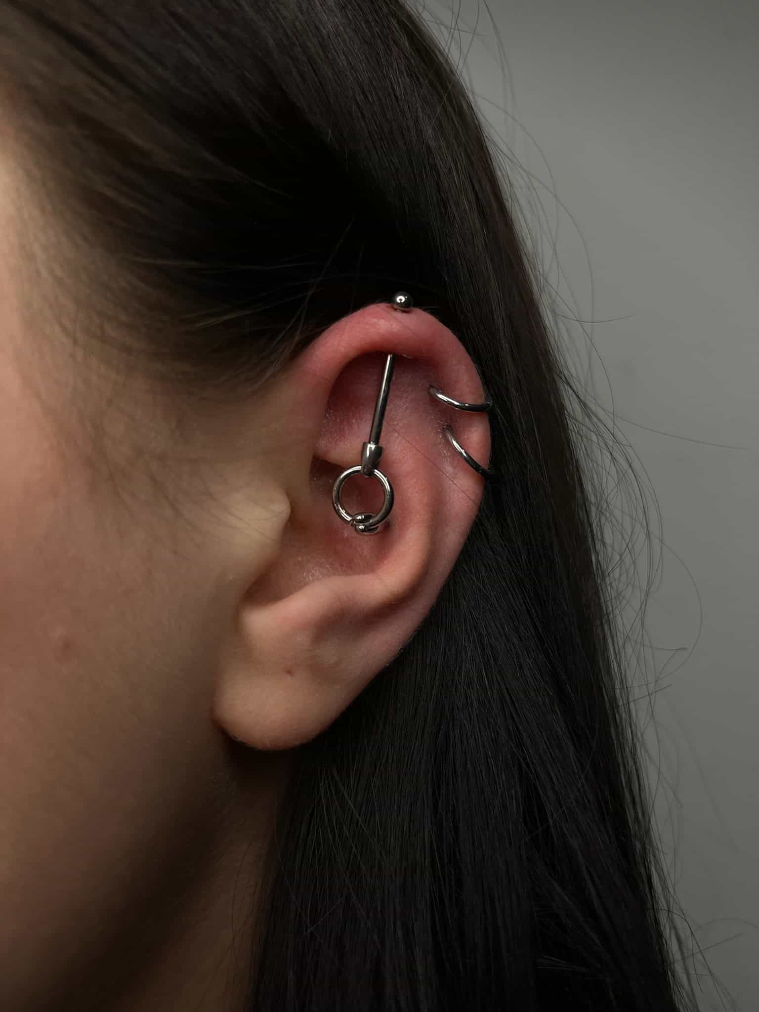 7sinstattoo.live Zbliżenie ucha ozdobionego różnymi piercingami, w tym małym kolczykiem kulkowym przy górnej krawędzi, kolczykiem w kształcie kółka otaczającym środek i industrialnym prętem przebijającym chrząstkę. Ucho wyróżnia się na neutralnym szarym tle, co jest przykładem kunsztu studia tatuażu Poznań. Ten obraz nosi tytuł IMG_4409. Najlepsze studio tatuażu Seven Sins Tattoo