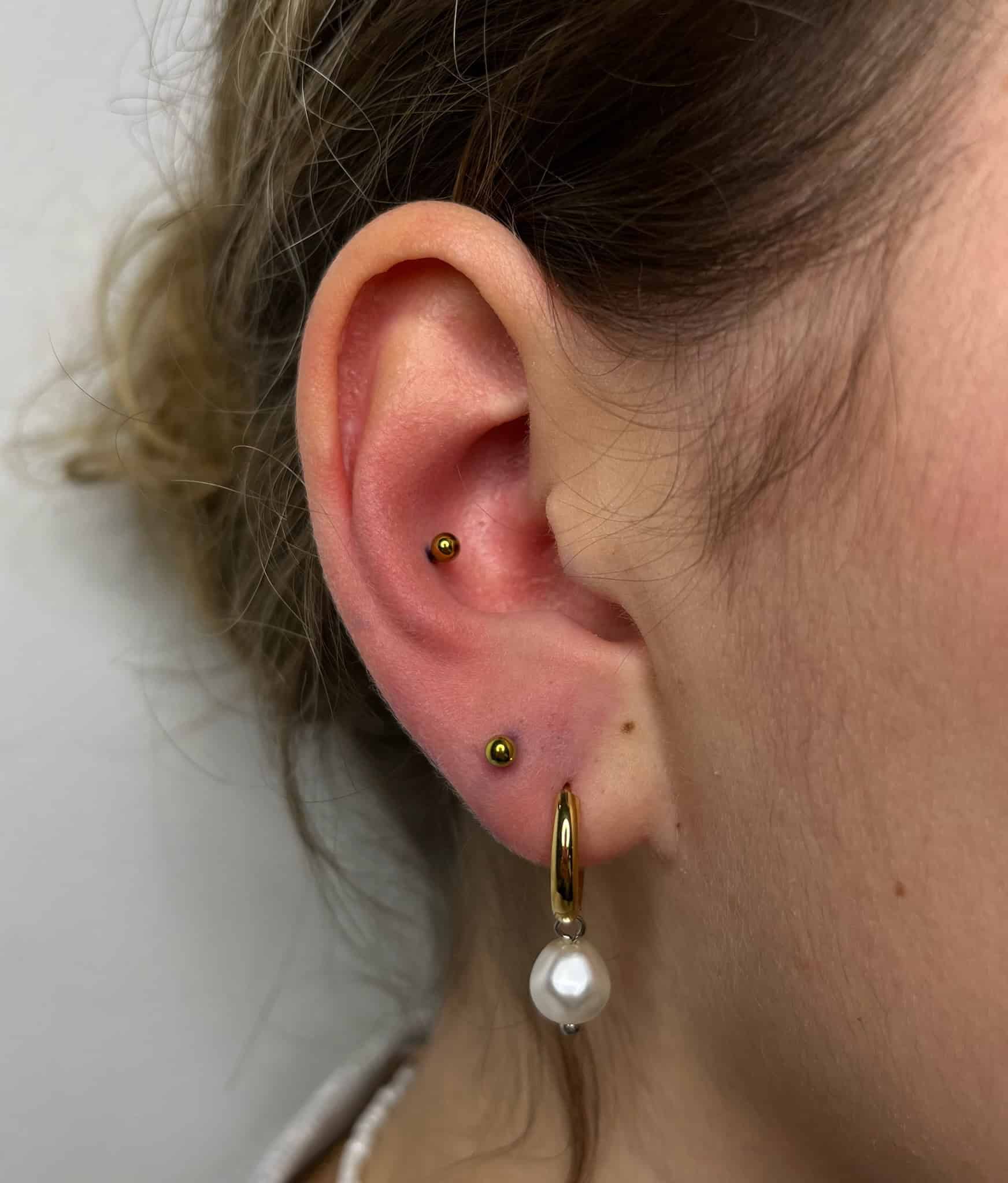 7sinstattoo.live Nieskazitelna fotografia oznaczona jako IMG_4849 ze studia tatuażu Poznań uchwyca z bliska ucho osoby ozdobione dwoma małymi złotymi kolczykami w górnej chrząstce i większym złotym kolczykiem w kształcie koła z wiszącą perłą w płatku ucha. Włosy są starannie zaczesane do tyłu, eksponując całe ucho na prostym i neutralnym tle. Najlepsze studio tatuażu Seven Sins Tattoo