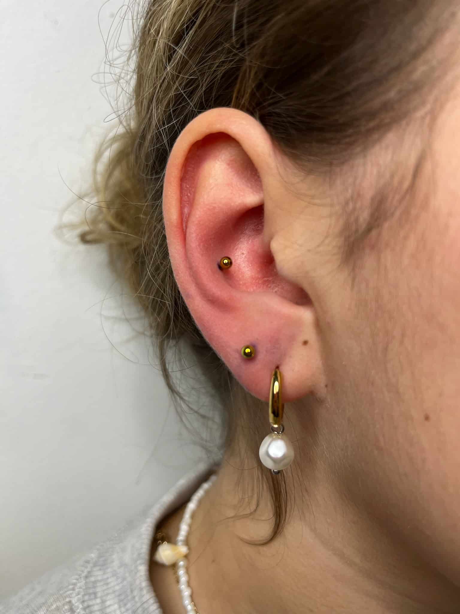 7sinstattoo.live Zbliżenie na ucho osoby z dwoma delikatnymi złotymi piercingami w okolicy helisy, któremu towarzyszy złoty kolczyk-kołnierz ozdobiony białą perłą na płatku. Ten wyrafinowany wygląd został sfotografowany w najlepszym studio tatuażu Poznań, znanym ze swojego kunsztu i precyzji. Osoba ma jasną skórę i związane blond włosy. Uchwycony obraz produktu jest oznaczony jako IMG_4851. Najlepsze studio tatuażu Seven Sins Tattoo