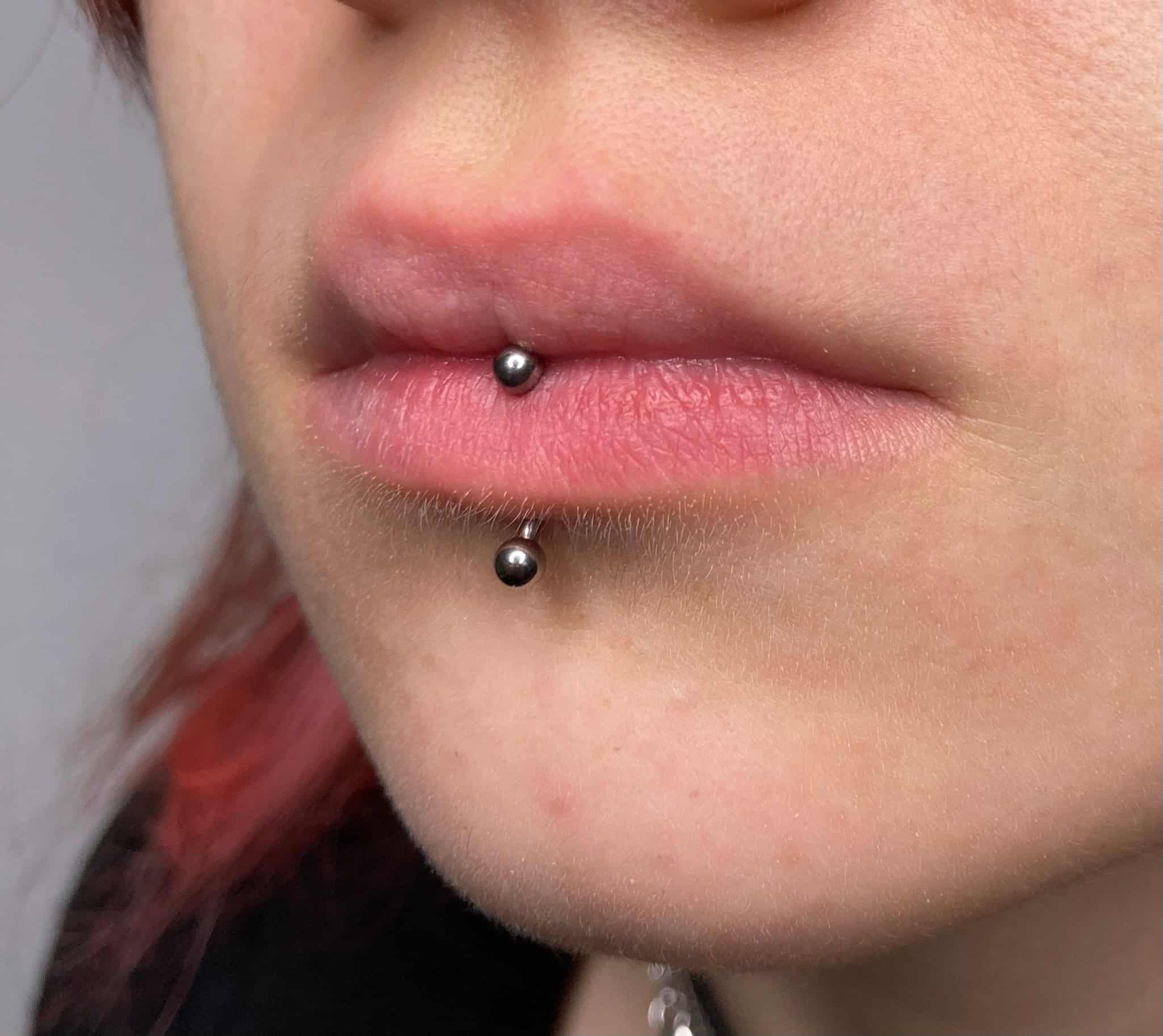 7sinstattoo.live Zbliżenie ust osoby pokazujące podwójne pionowe piercingi wargi górnej. Górny piercing znajduje się na dole dolnej wargi, a dolny piercing tuż nad brodą. Oba piercingi mają małą, okrągłą metalową biżuterię. Zdjęcie, zidentyfikowane jako IMG_4897, zostało zrobione w renomowanym studiu tatuaży Poznań na neutralnym tle. Najlepsze studio tatuażu Seven Sins Tattoo