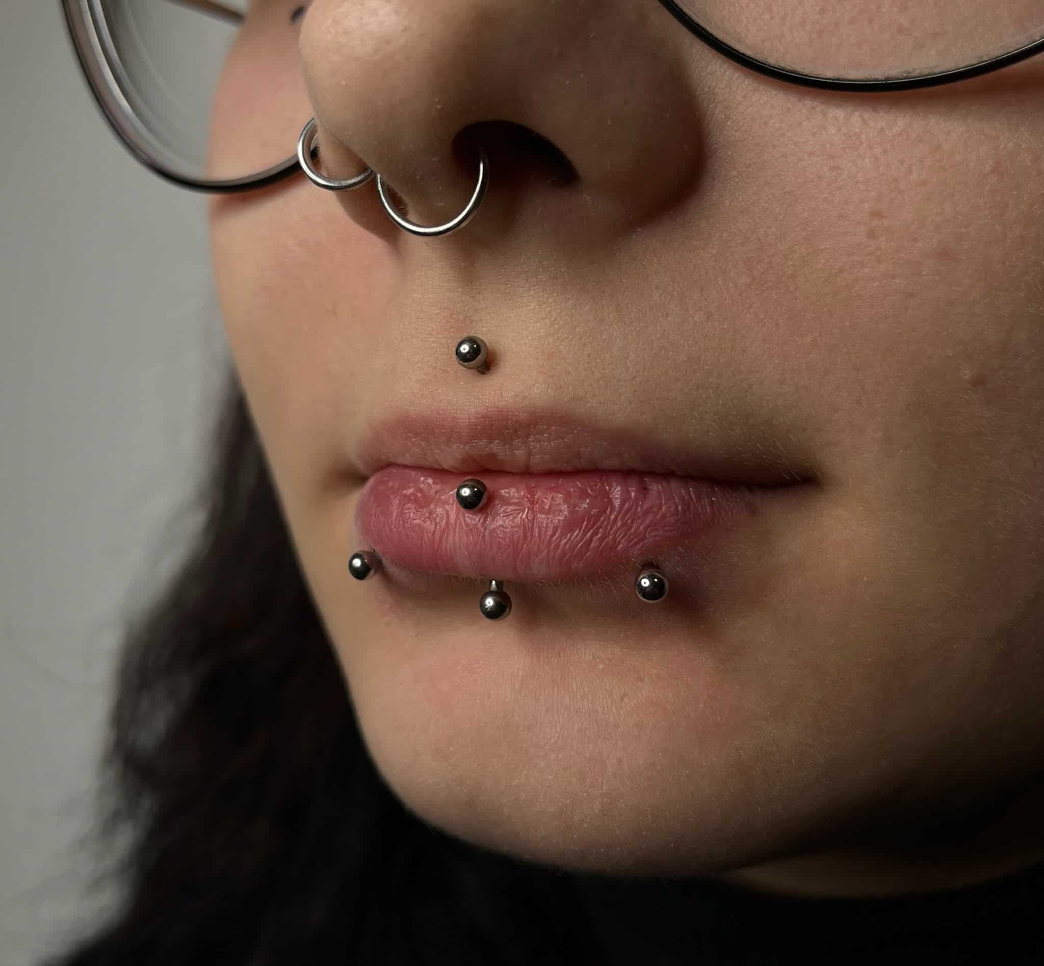 7sinstattoo.live Zbliżenie osoby noszącej okulary, z piercingiem twarzy. Piercing obejmuje kółko w przegrodzie nosowej, kolczyk w wargę górną i poziomy rząd trzech kolczyków pod dolną wargą. Uchwycony w studio tatuażu Poznań, IMG_5168 przedstawia osobę z neutralnym wyrazem twarzy. Najlepsze studio tatuażu Seven Sins Tattoo
