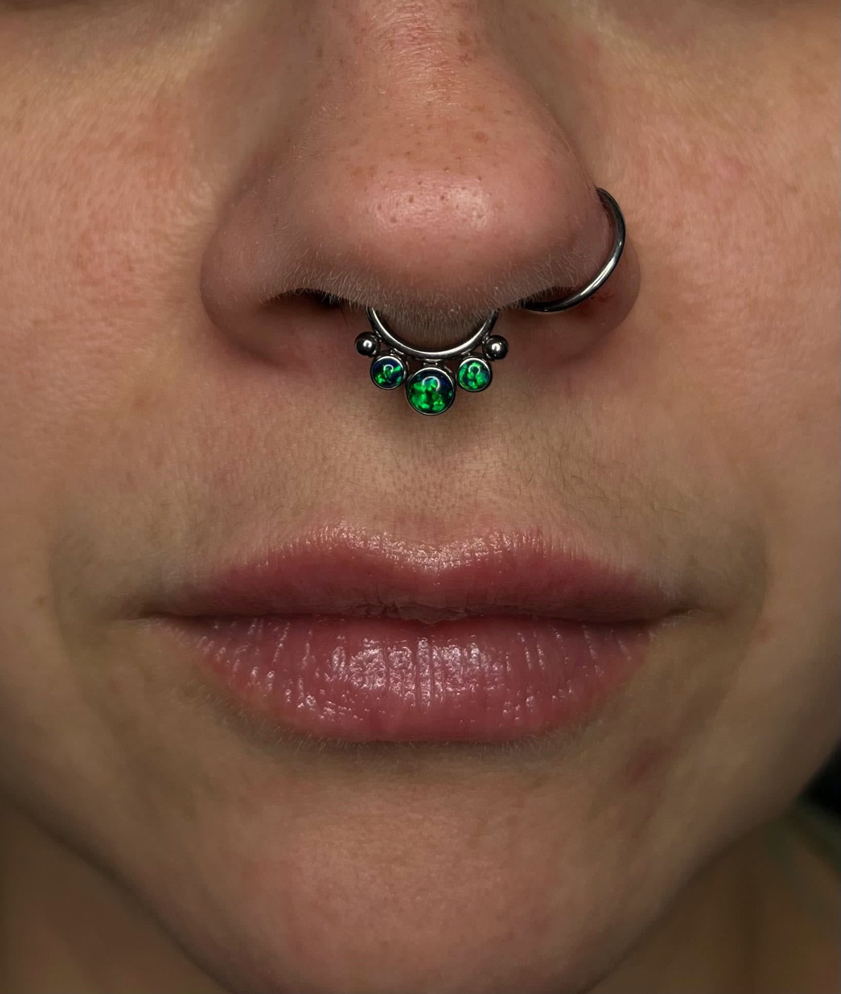 7sinstattoo.live Zbliżenie z najlepszego studia tatuażu Poznań pokazuje usta i nos osoby z piercingiem przegrody nosowej. Okrągły sztanga, nazwany IMG_5173, jest ozdobiony trzema zielonymi klejnotami i znajduje się tuż pod nosem, podczas gdy odrobina balsamu do ust lub błyszczyka dodaje subtelnego połysku. Najlepsze studio tatuażu Seven Sins Tattoo