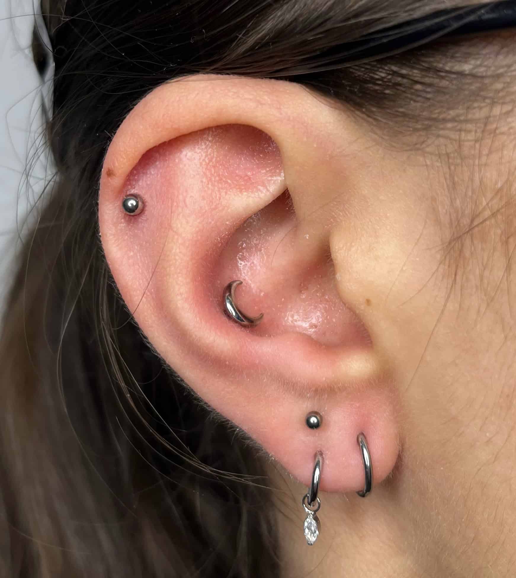 7sinstattoo.live Zbliżenie ucha osoby z czterema piercingami: mały kolczyk na górnej chrząstce, mały kółko w uchu wewnętrznym, kolczyk na płatku ucha i dwa kółka na dolnej zewnętrznej krawędzi ucha, jeden z małym zwisającym kryształkiem. Osoba ma na sobie okulary w ciemnej oprawie ze studia tatuażu Poznań. Ten obraz nosi tytuł IMG_5618. Najlepsze studio tatuażu Seven Sins Tattoo