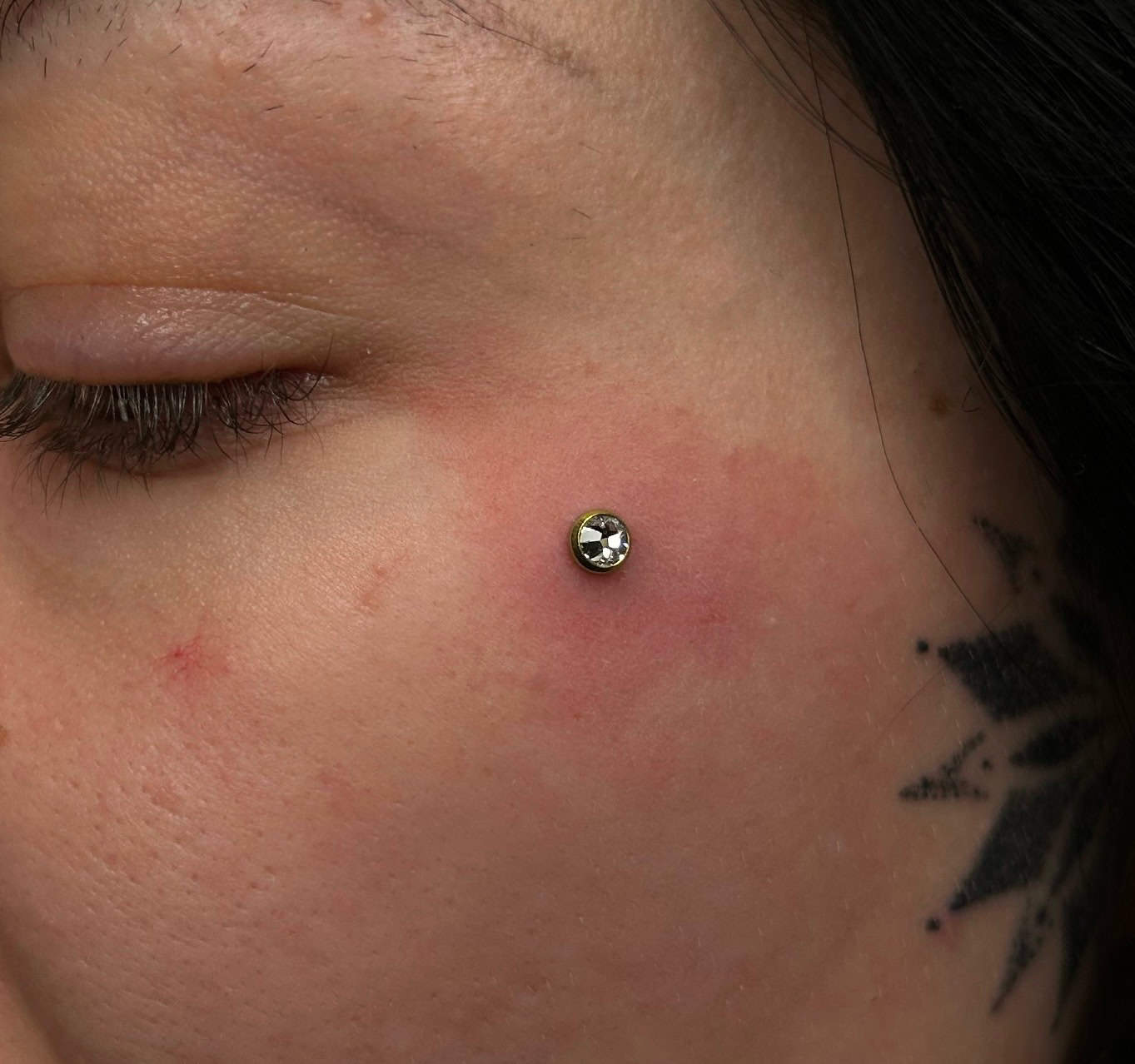 7sinstattoo.live Zbliżenie na policzek osoby pokazujące IMG_5683, piercing skórny z okrągłym, srebrnym klejnotem. Skóra wokół piercingu jest lekko zaczerwieniona. Część czarnego geometrycznego tatuażu jest widoczna w pobliżu piercingu, prezentując kunszt studia tatuaży Poznań, a włosy osoby są ciemne. Oko osoby jest zamknięte. Najlepsze studio tatuażu Seven Sins Tattoo
