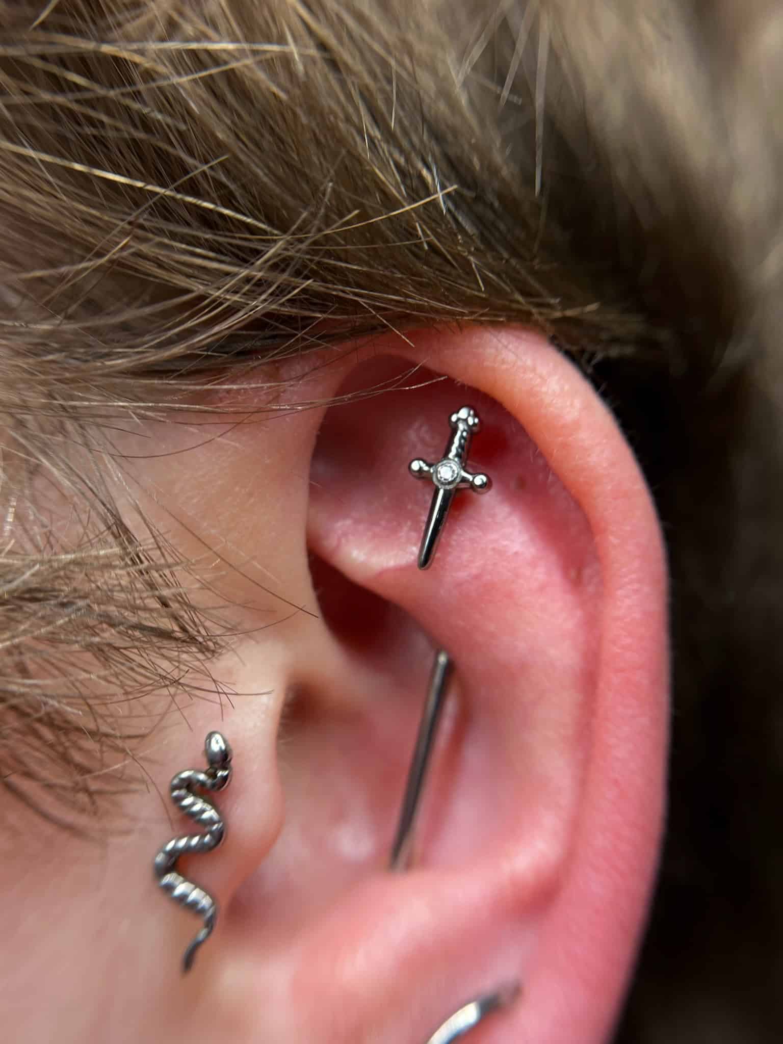 7sinstattoo.live Zbliżenie ucha z trzema piercingami zaprezentowanymi w najlepszym studio tatuażu w Poznaniu. Piercingi obejmują mały kolczyk w kształcie miecza w chrząstce, srebrny kolczyk w kształcie sztangi nad nim i kolczyk w kształcie węża w płatku ucha. Jasne włosy są częściowo widoczne w tle. Odniesienie do obrazu: IMG_6075. Najlepsze studio tatuażu Seven Sins Tattoo