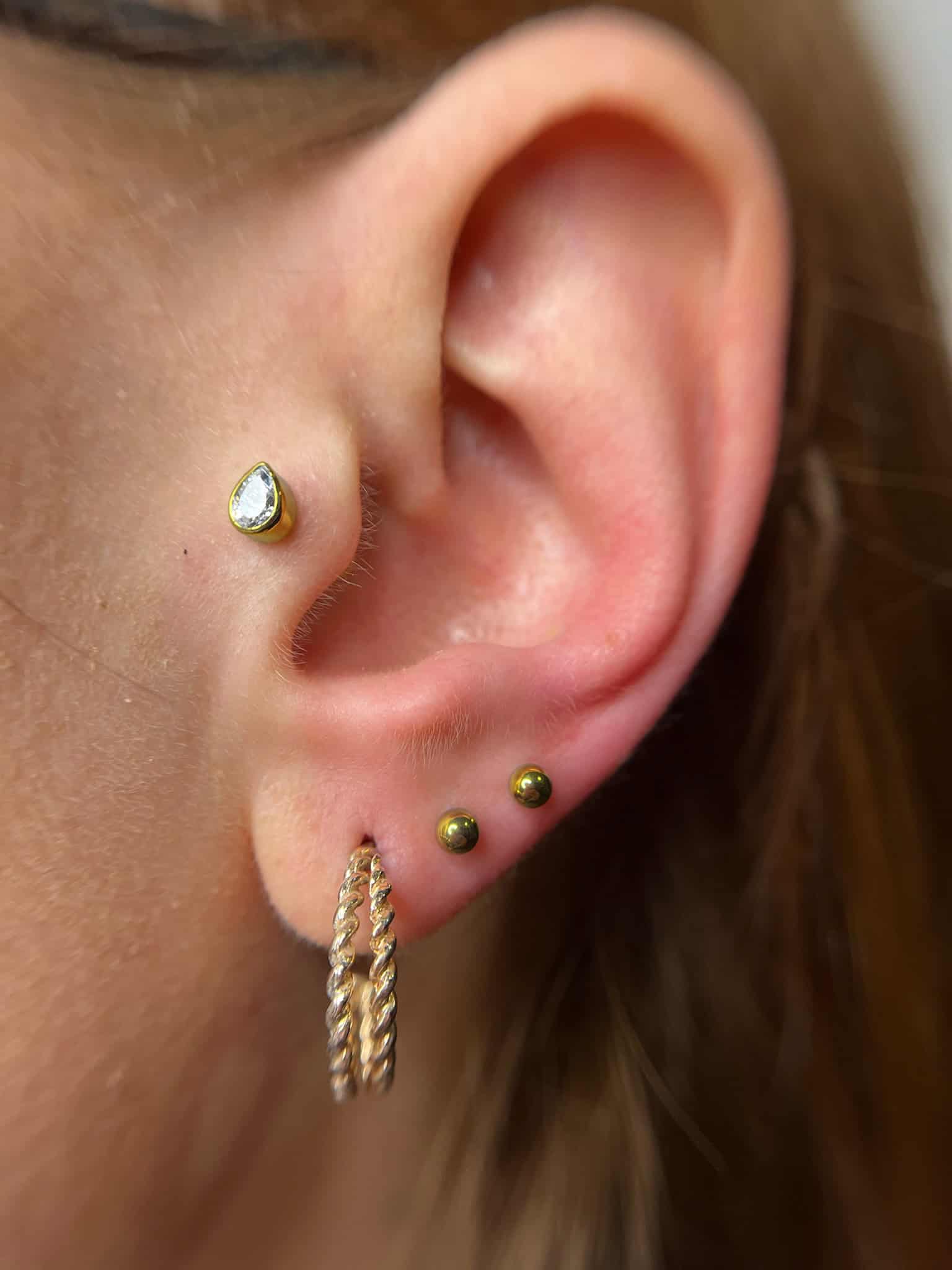 7sinstattoo.live Zbliżenie ucha ozdobionego wieloma piercingami ze studia tatuaży Poznań. Ucho ma mały, przezroczysty kolczyk z kamieniem szlachetnym u góry, dwa małe złote kolczyki kulkowe na płatku ucha i złoty kolczyk-kółko ze skręconym wzorem poniżej płatka ucha, uchwycony w produkcie IMG_6132. Najlepsze studio tatuażu Seven Sins Tattoo