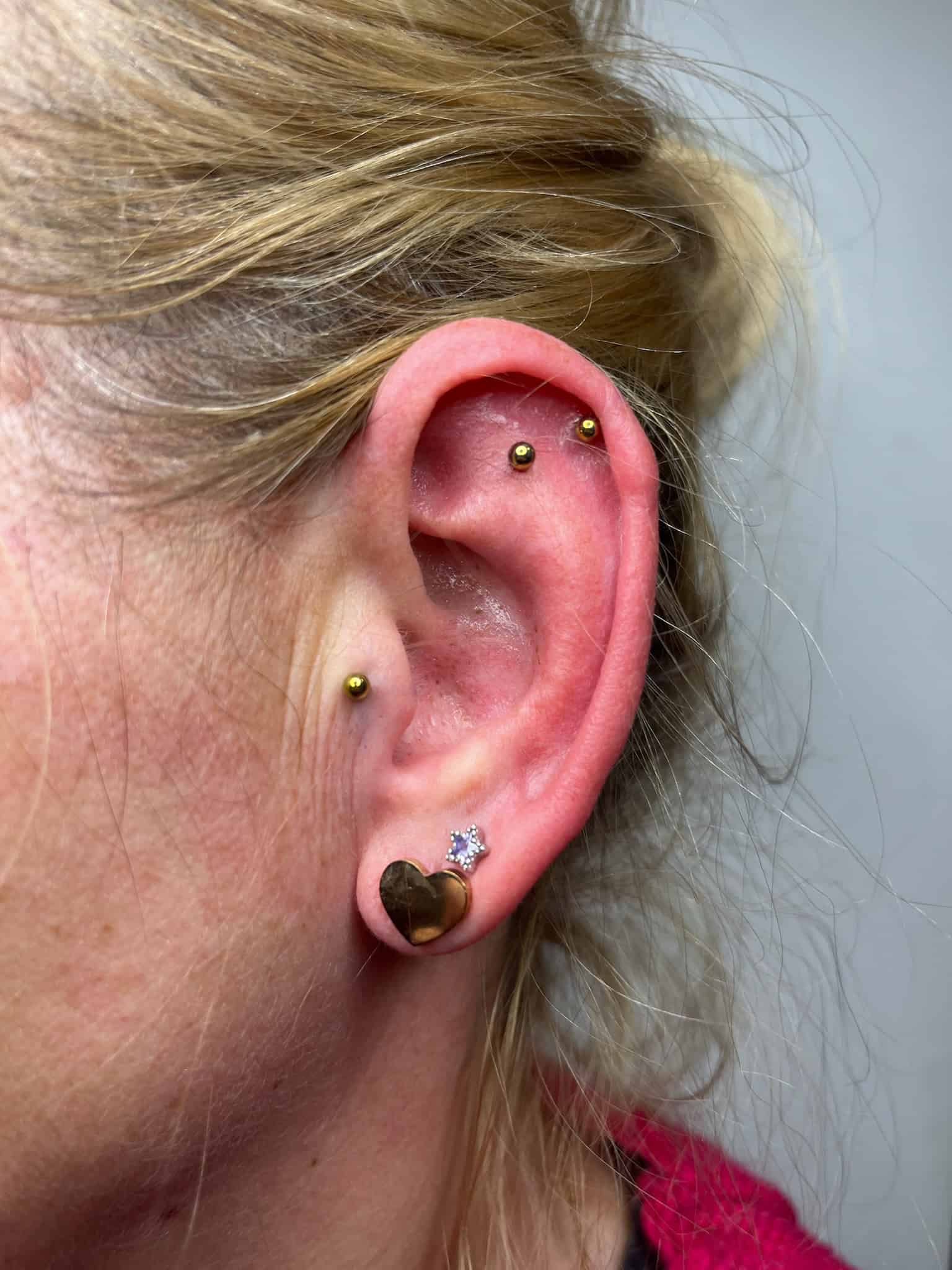 7sinstattoo.live Zbliżenie na zewnętrzne ucho osoby z wieloma piercingami, zrobione w studio tatuażu Poznań. Ucho prezentuje obraz IMG_6205, ze złotym kolczykiem na płatku, jednym złotym piercingiem z klejnotem w kształcie gwiazdy i dwoma złotymi okrągłymi sztangami na górnej części. Osoba ma blond włosy związane z tyłu i nosi czerwony strój. Najlepsze studio tatuażu Seven Sins Tattoo