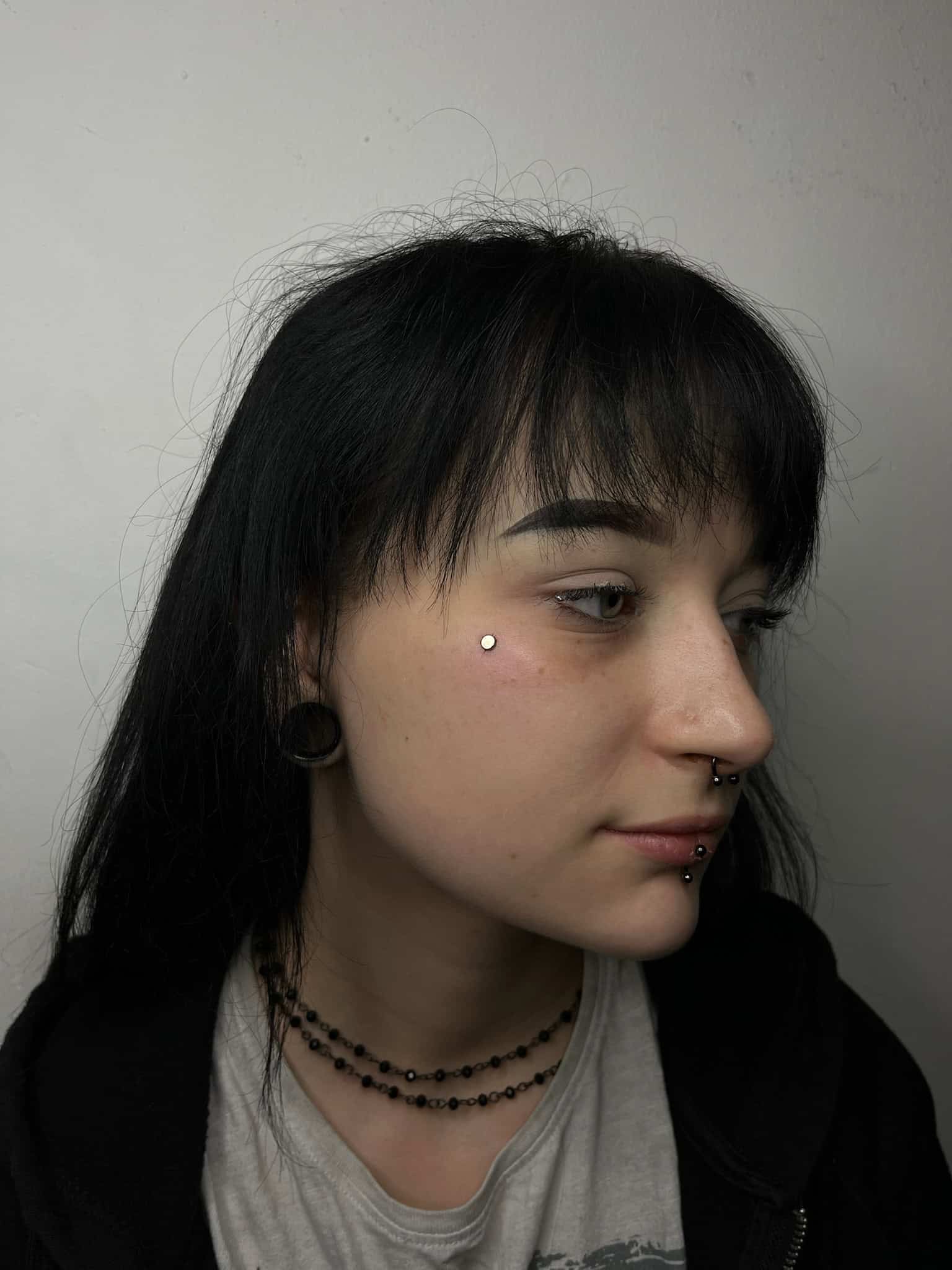 7sinstattoo.live Na zdjęciu zatytułowanym IMG_6351 osoba z prostymi, ciemnymi włosami i grzywką jest ozdobiona wieloma piercingami twarzy, w tym kolczykiem na policzku, kolczykiem w przegrodzie nosowej i biżuterią w ustach. Ma na sobie czarny naszyjnik z koralików i czarną bluzę z kapturem na jasnej koszuli na jednolitym tle w studio tatuażu Poznań. Najlepsze studio tatuażu Seven Sins Tattoo