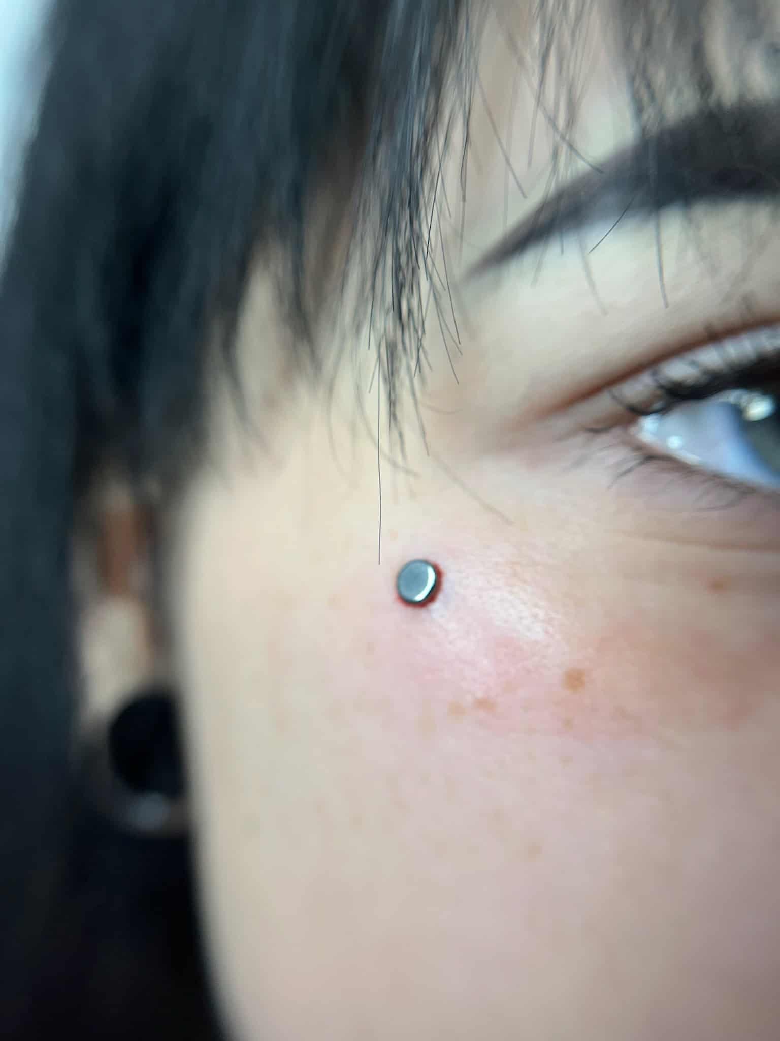 7sinstattoo.live Zbliżenie prawej strony twarzy osoby, podkreślające nowy piercing skórny w pobliżu kości policzkowej od *studio tatuażu Poznań*. Piercing prezentuje mały, okrągły, srebrny kolczyk. Widoczne jest oko osoby i część jej brwi, z lekkim rumieńcem na policzku. Nazwa produktu: IMG_6356. Najlepsze studio tatuażu Seven Sins Tattoo