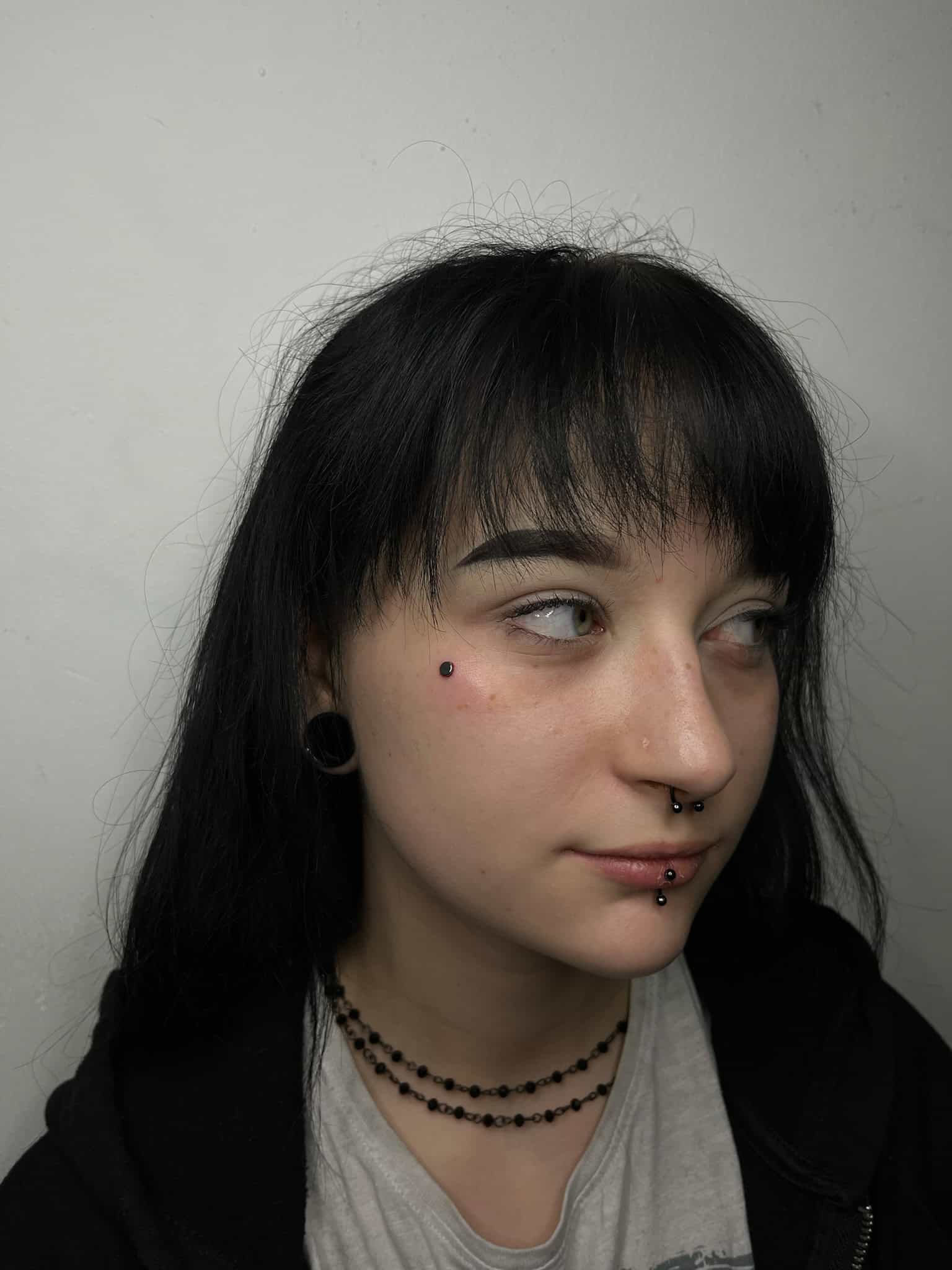 7sinstattoo.live IMG_6365 przedstawia osobę z długimi czarnymi włosami, ciemną grzywką i wieloma piercingami twarzy, w tym piercingiem brwi, nosa i warg. Patrzy w bok, mając na sobie czarny kaptur i czarne naszyjniki z koralików. Osoba ta zachowuje neutralny wyraz twarzy przed jednolitym tłem w studio tatuażu Poznań. Najlepsze studio tatuażu Seven Sins Tattoo