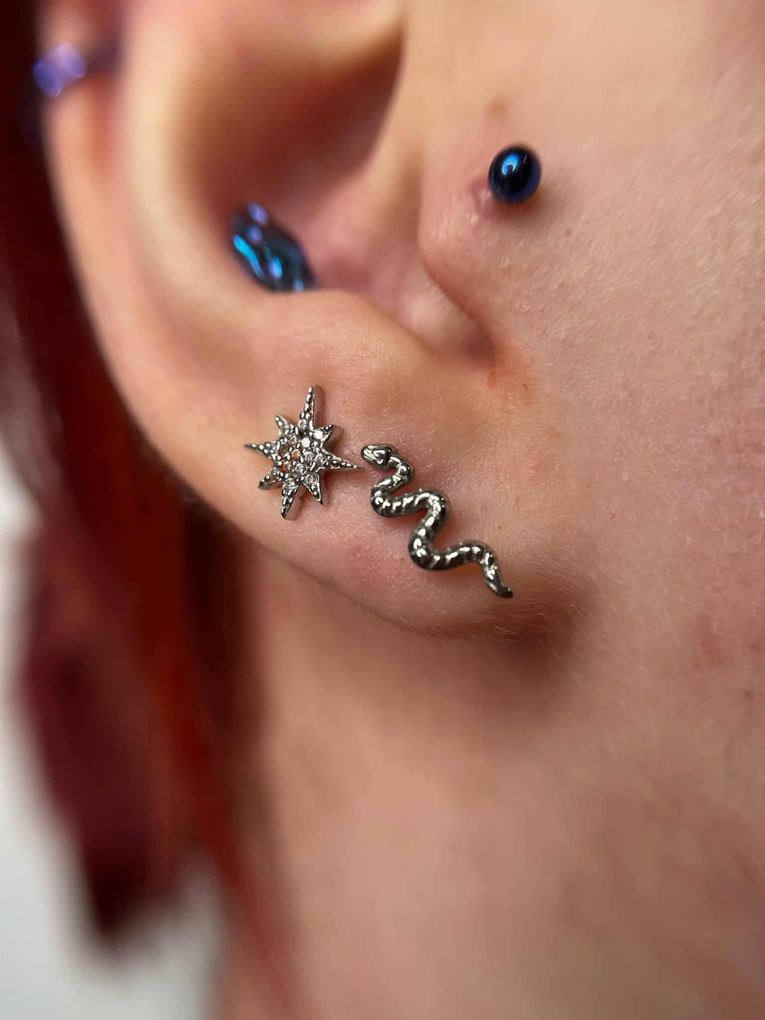 7sinstattoo.live Zbliżenie na ucho z dwoma piercingami: jeden kolczyk w chrząstce ozdobiony niebieskim klejnotem i piercing płatka ucha ze srebrnym kolczykiem w kształcie węża wysadzanym małymi klejnotami, wszystko wykonane w studio tatuaży Poznań. Dodatkowo, kolejny mały piercing chrząstki z niebieskim klejnotem znajduje się bliżej głowy. Obraz jest oznaczony jako IMG_6664. Najlepsze studio tatuażu Seven Sins Tattoo