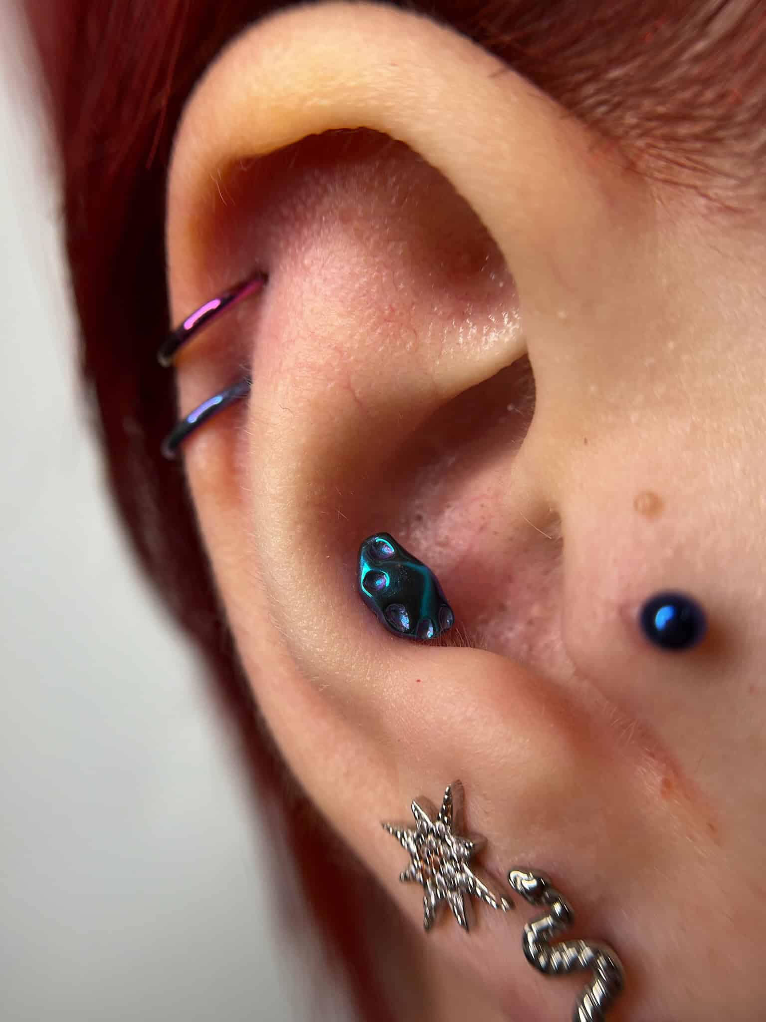7sinstattoo.live Zbliżenie ucha osoby ozdobionego wieloma kolczykami. Ucho ma pięć odrębnych elementów biżuterii, w tym dwa kółka u góry, mały niebieski kolczyk w środku ucha, kolczyk w kształcie gwiazdy na dolnej części i kolczyk w kształcie węża na dole — idealny do pokazania się po wizycie w tatuaż Poznań. Nazwa produktu: IMG_6668. Najlepsze studio tatuażu Seven Sins Tattoo